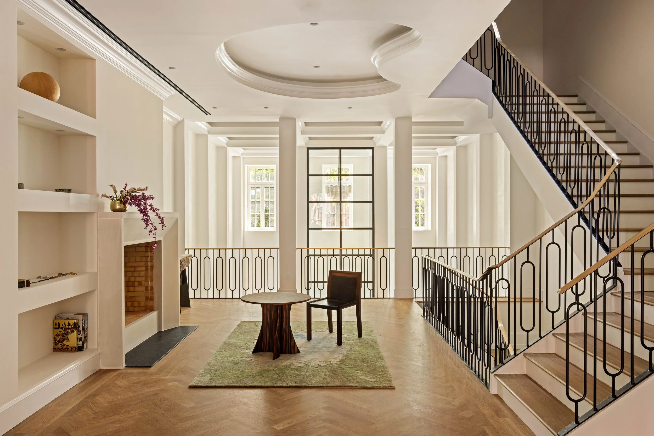 Bright foyer with high ceilings, large windows, wooden floors, a small round table, and two chairs, featuring a staircase with wrought iron railings and decorative molding.