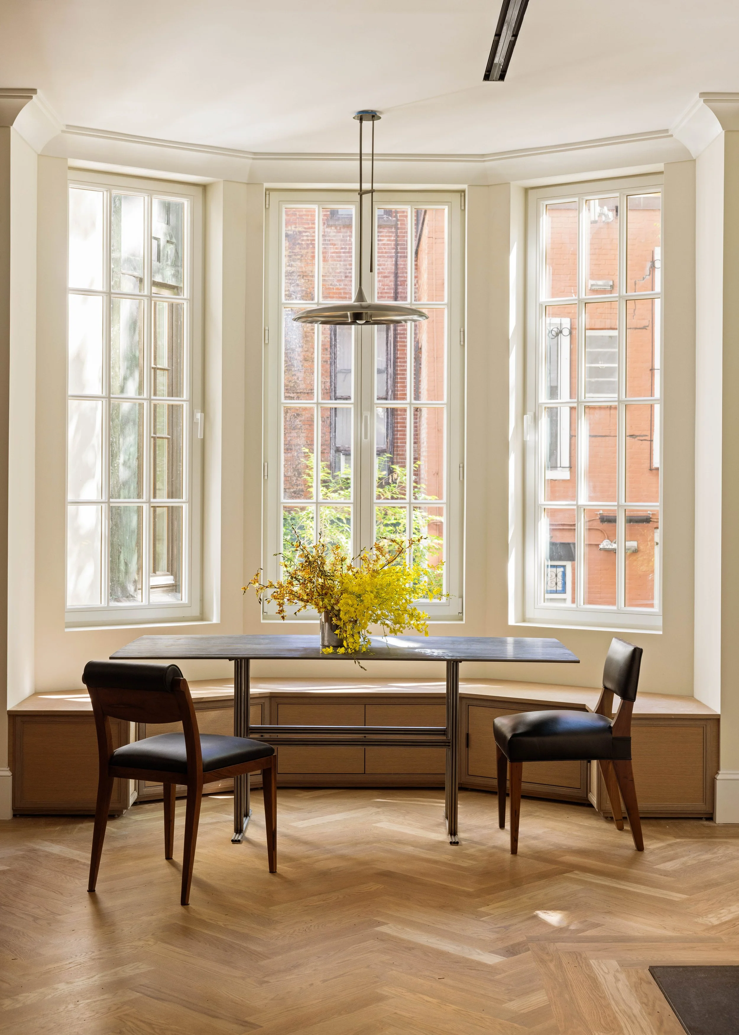 A dining area with a wooden table, three black chairs, and a large window with a view of brick buildings outside. A yellow flower arrangement decorates the table, and a modern pendant light hangs from the ceiling.