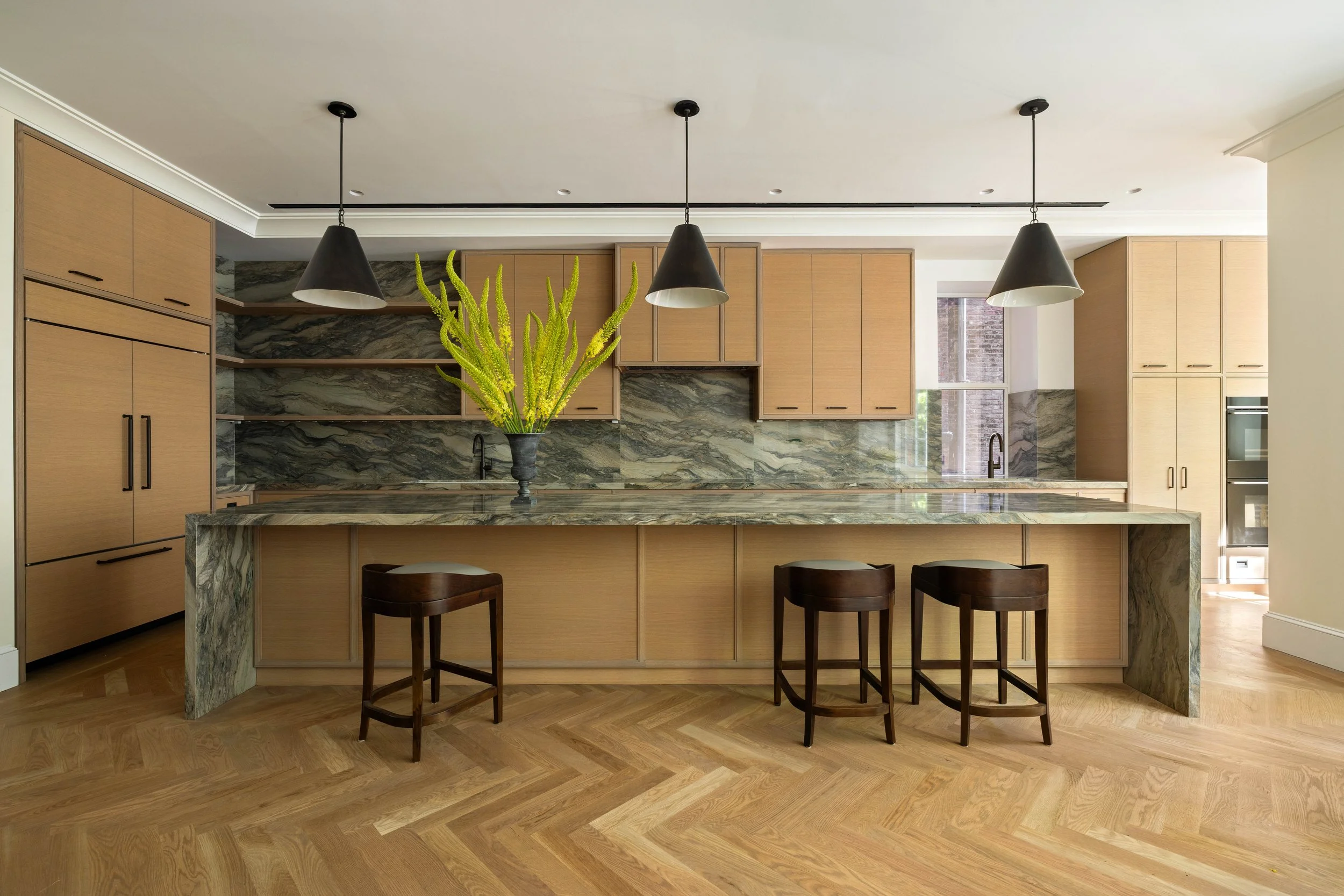 Modern kitchen with a marble island, light wood cabinets, black hardware, black pendant lights, a green plant in a black vase, and large windows with a view outside.