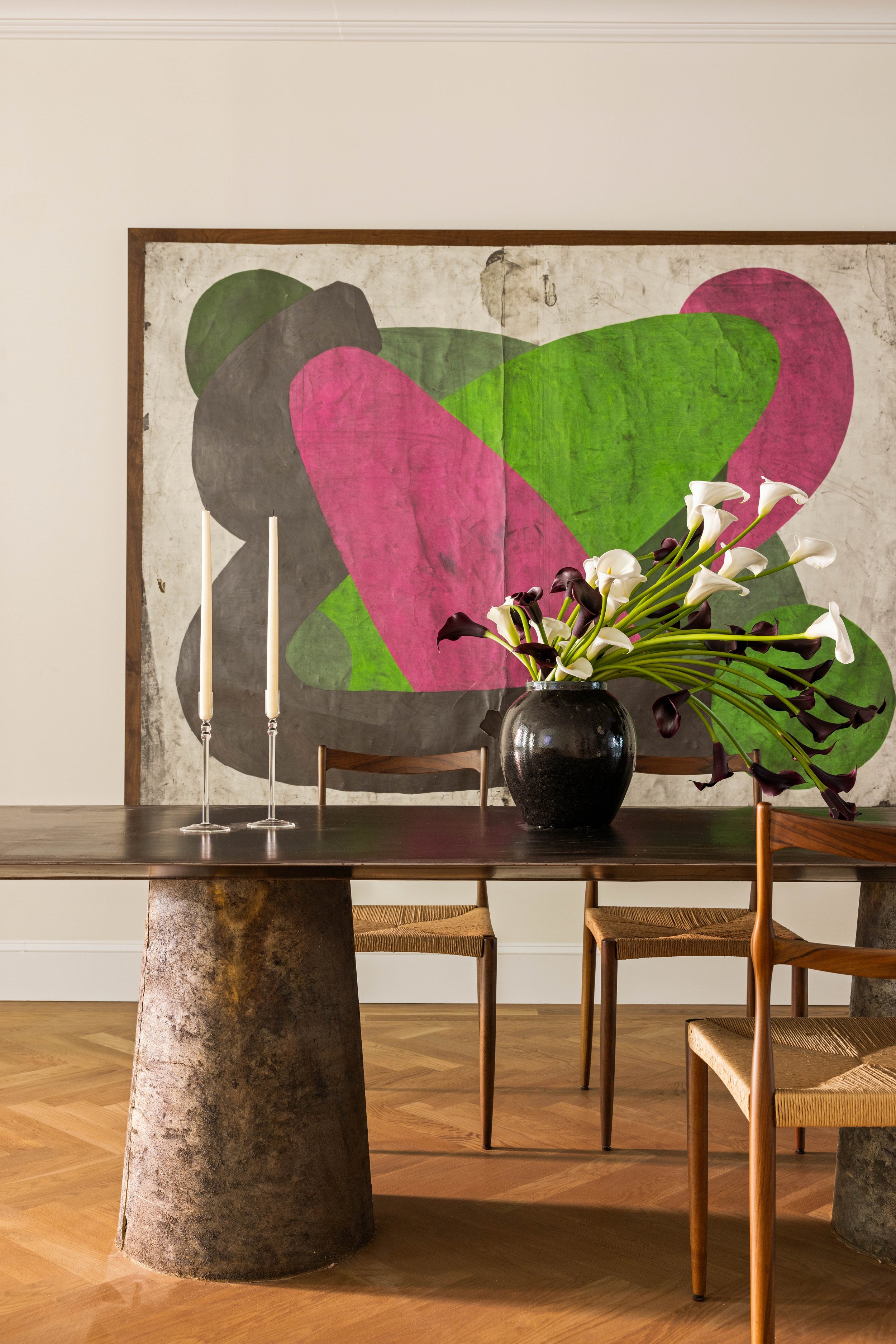 A dining room with a wooden table, four wooden chairs with woven seats, two white candlesticks, a black vase with white and dark purple calla lilies, and a large abstract painting with pink, green, black, and beige shapes on the wall