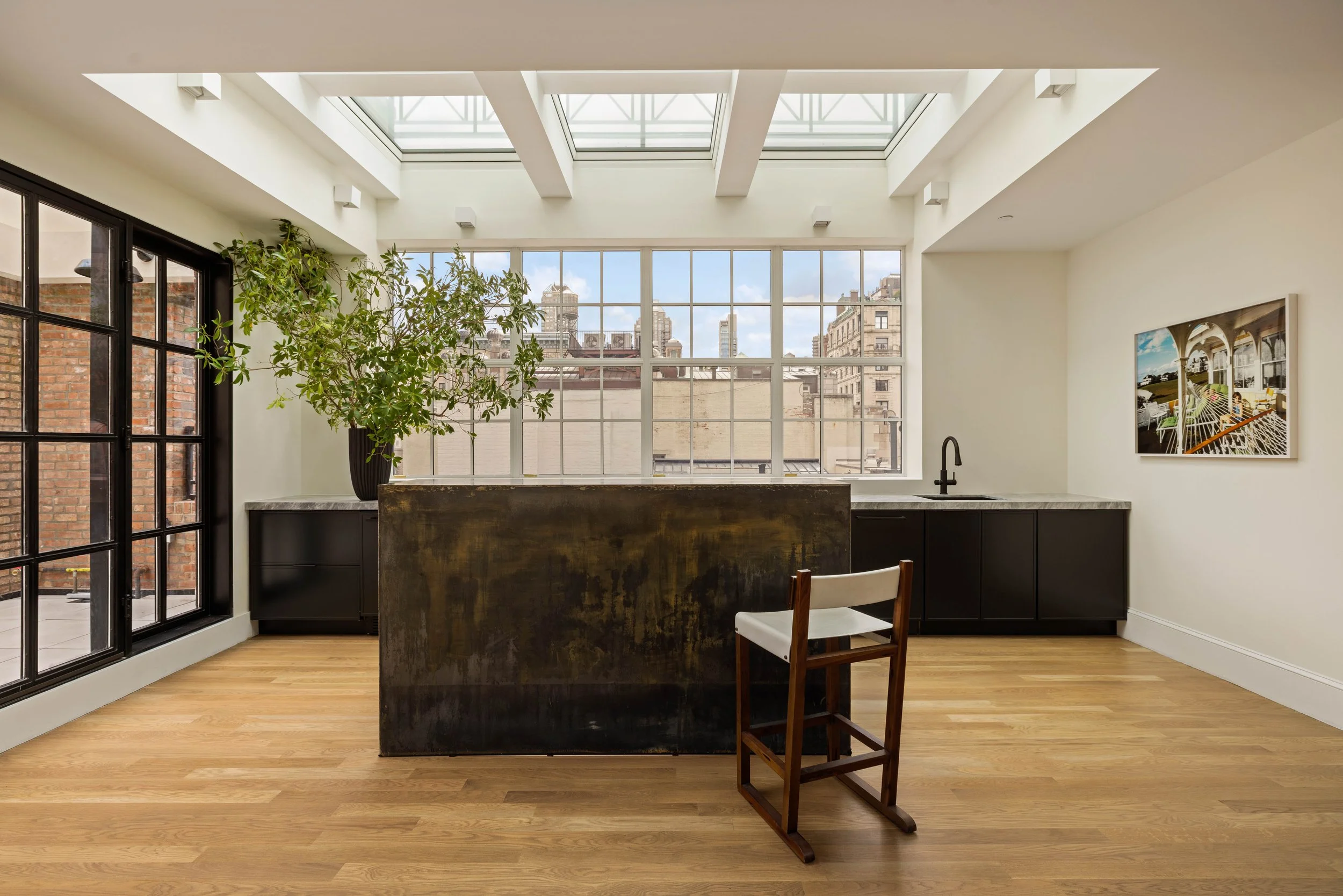 Modern kitchen with large windows, skylights, wooden floor, black cabinetry, a large potted plant, and a piece of colorful artwork on the wall.