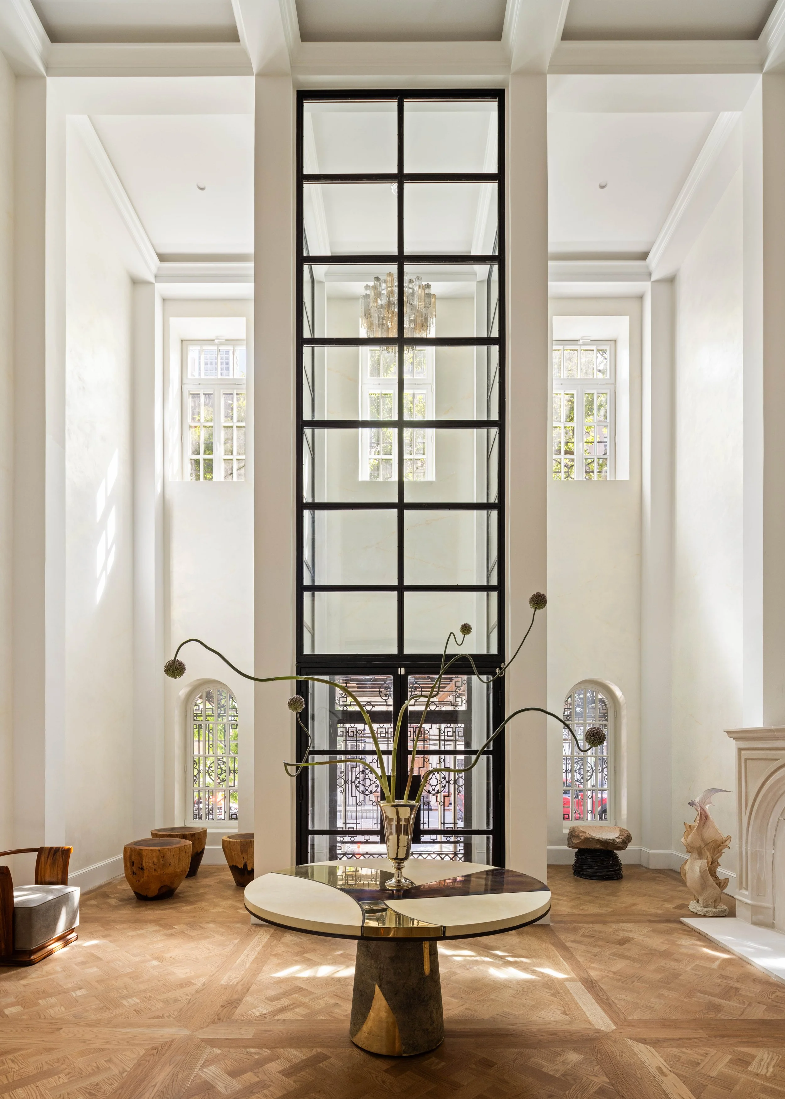 Interior of a spacious, elegant room with high ceilings and large windows, featuring a central black-framed glass door or window, a round table with a decorative vase and flowers, wooden stools, sculptures, and natural light.