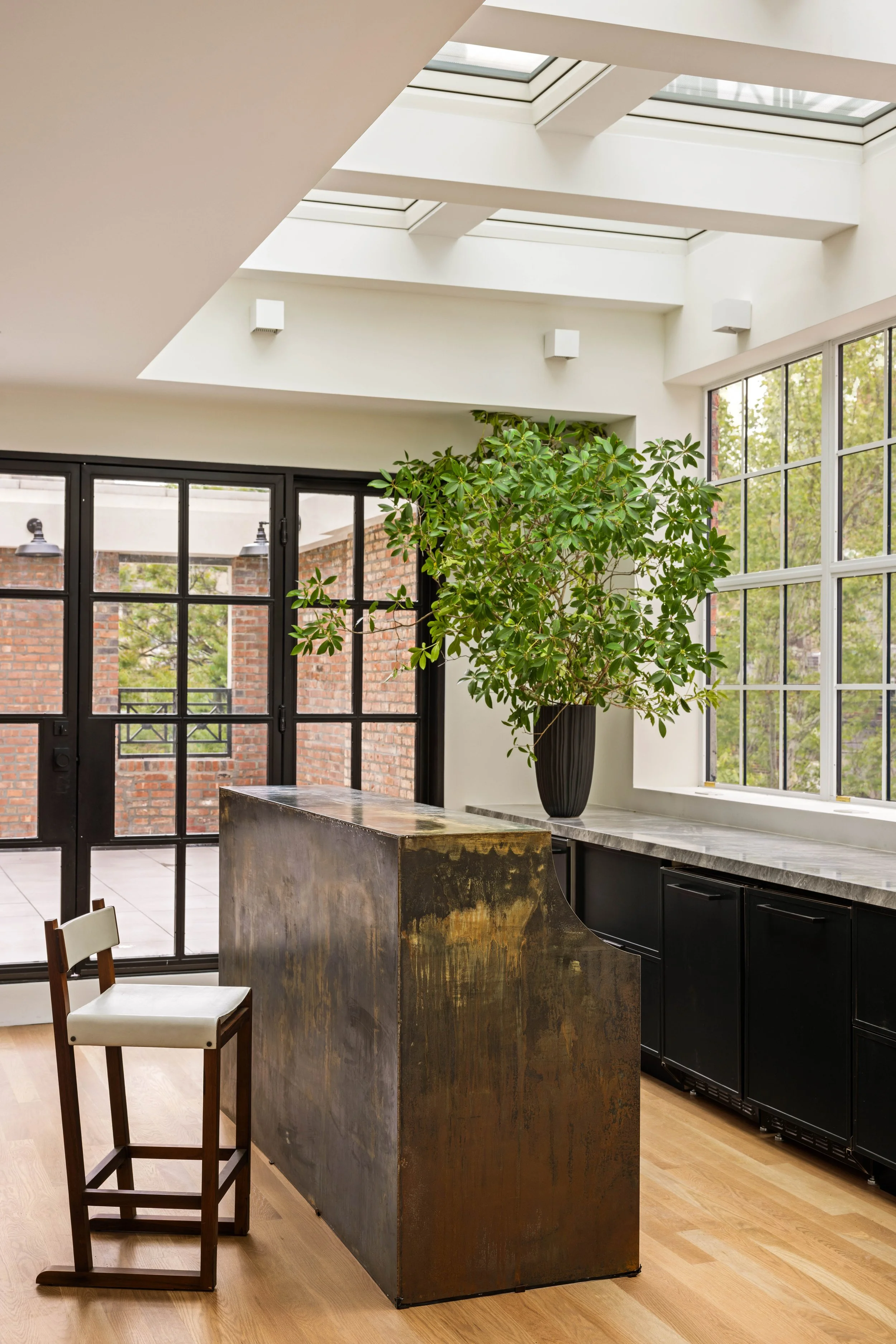 Interior of a modern kitchen with large windows, a potted plant, and a rustic kitchen island made of metal with a wood-like finish. There is a single chair with a white cushion and dark wood frame. Outside views show brick buildings and trees.