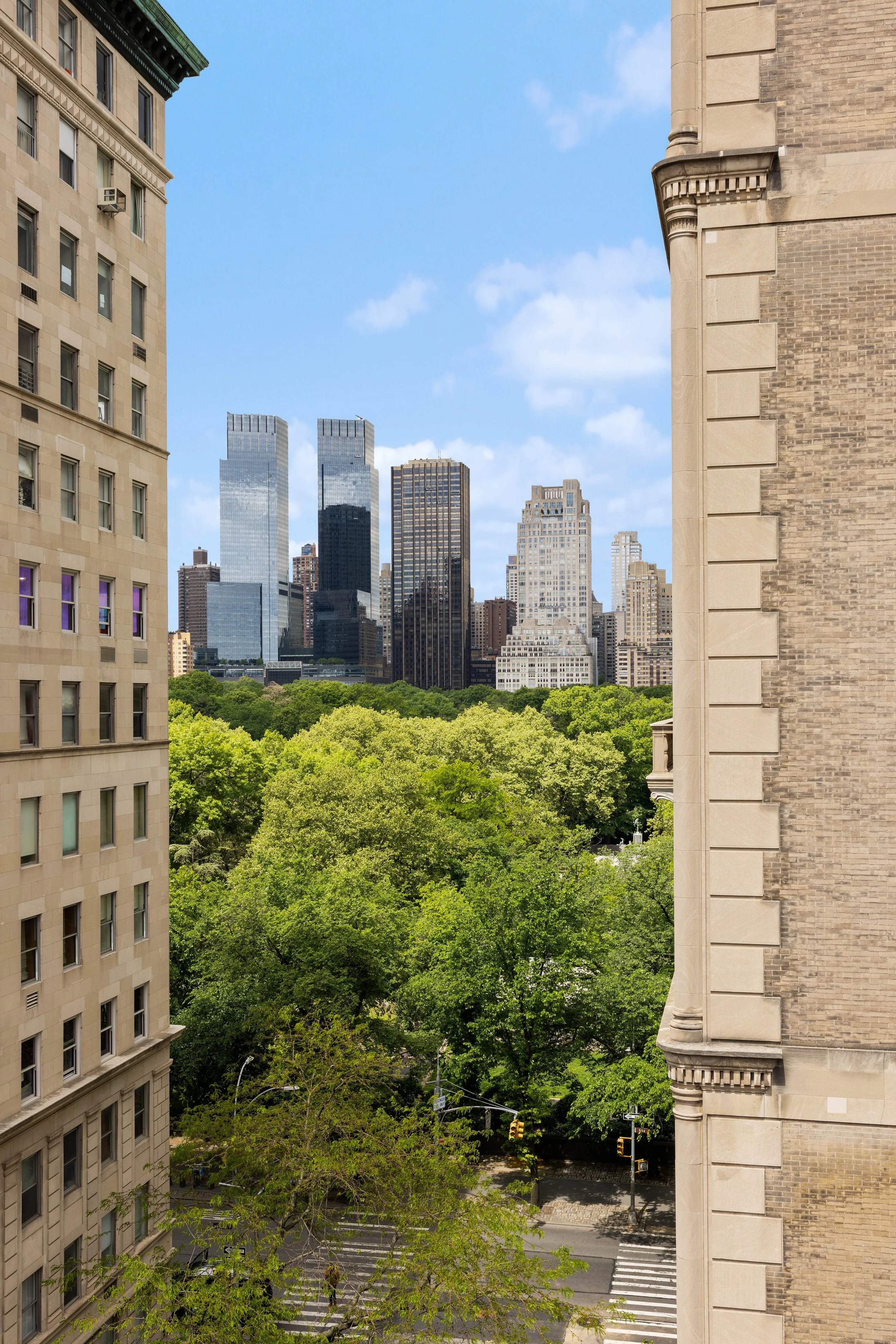 View of a city skyline with tall modern glass buildings in the background, lush green trees in the foreground, and older brick and stone buildings on the sides, under a partly cloudy blue sky.
