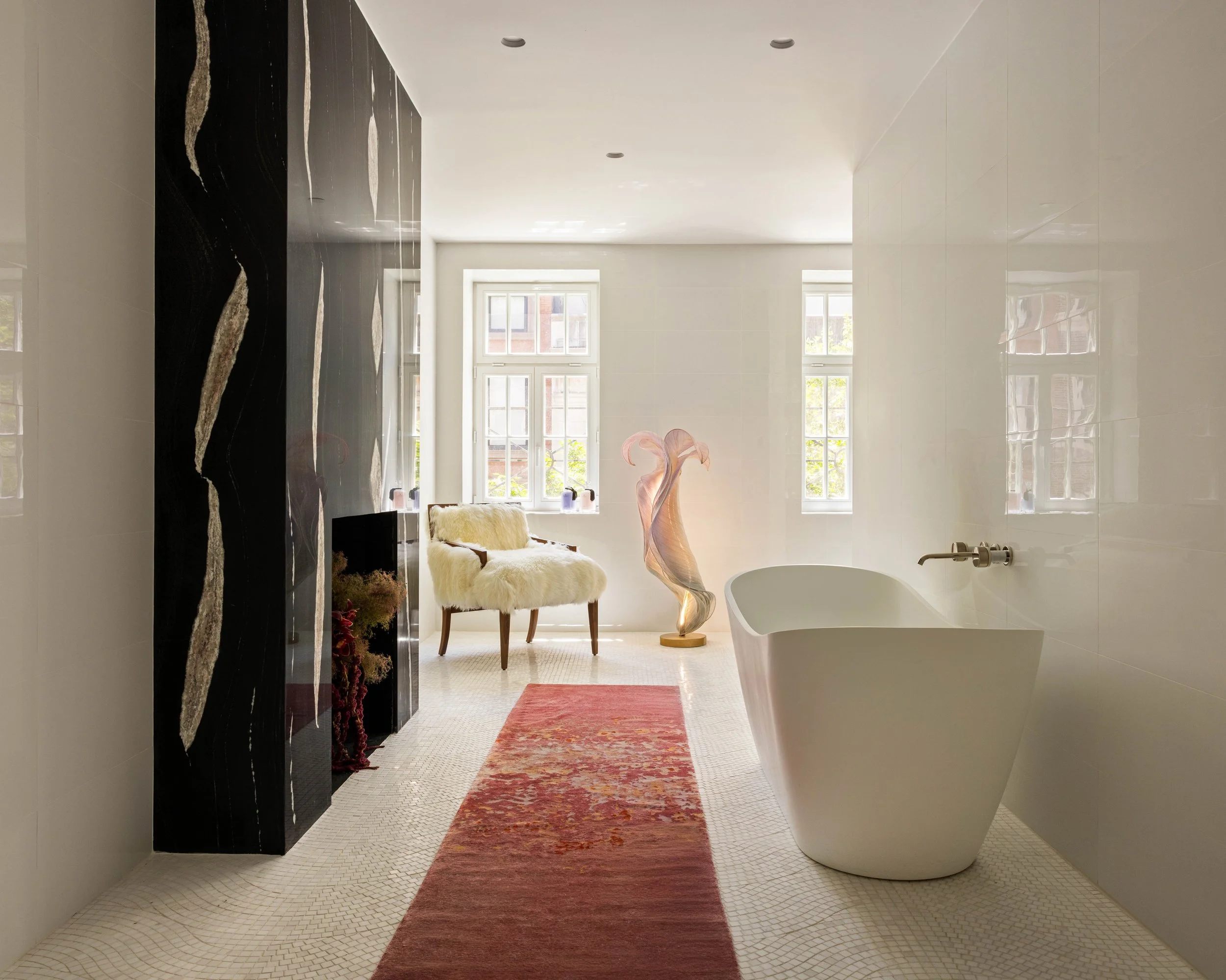 A bathroom with a white freestanding bathtub, an ornate pink glass sculpture, a plush white armchair with a furry throw, and large windows providing natural light. The floor has small white tiles with a red and orange patterned rug.