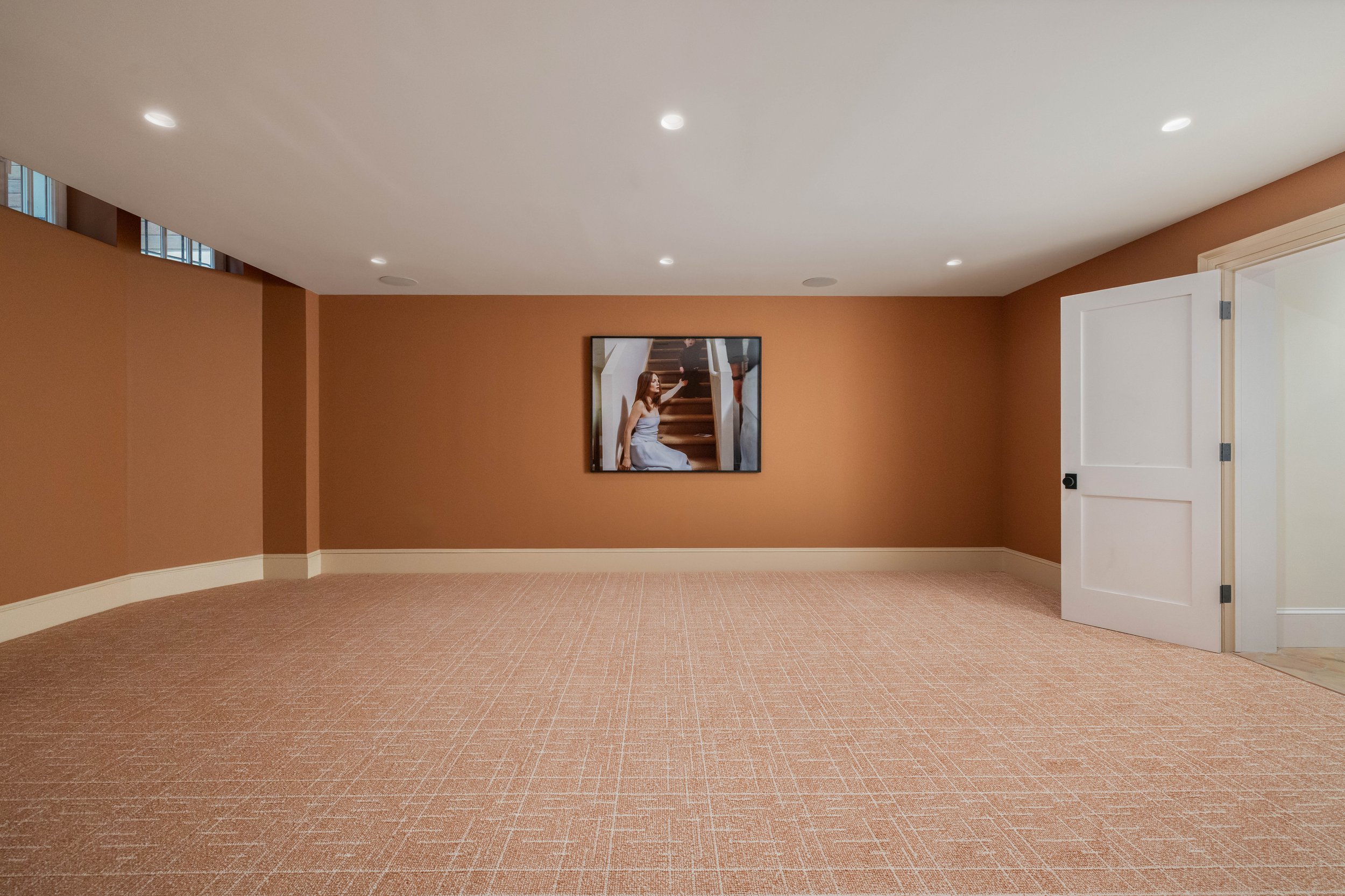 Empty room with beige carpet, brown walls, ceiling lights, a white door open to another room, and a framed photo of a woman on stairs.