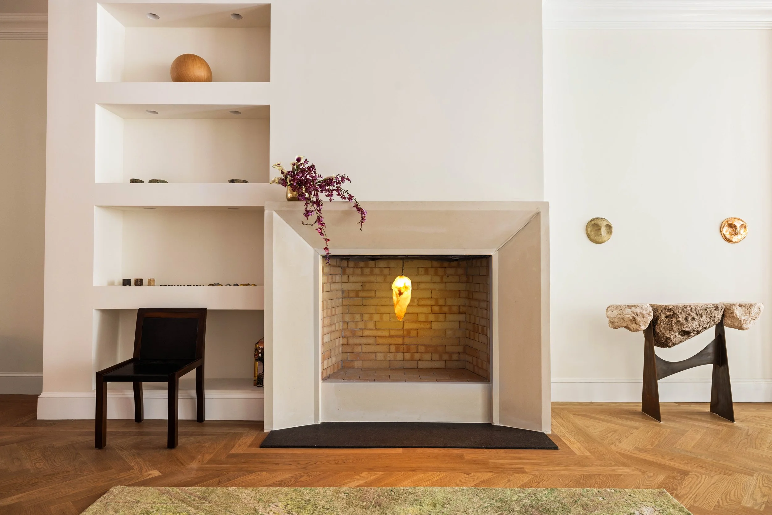 Living room with white built-in shelves, a fireplace with brick interior, a hanging light, and minimalist decor including a decorative vase with purple flowers, ceramic objects on the shelves, a black chair, and a stone sculpture on a wooden stand.