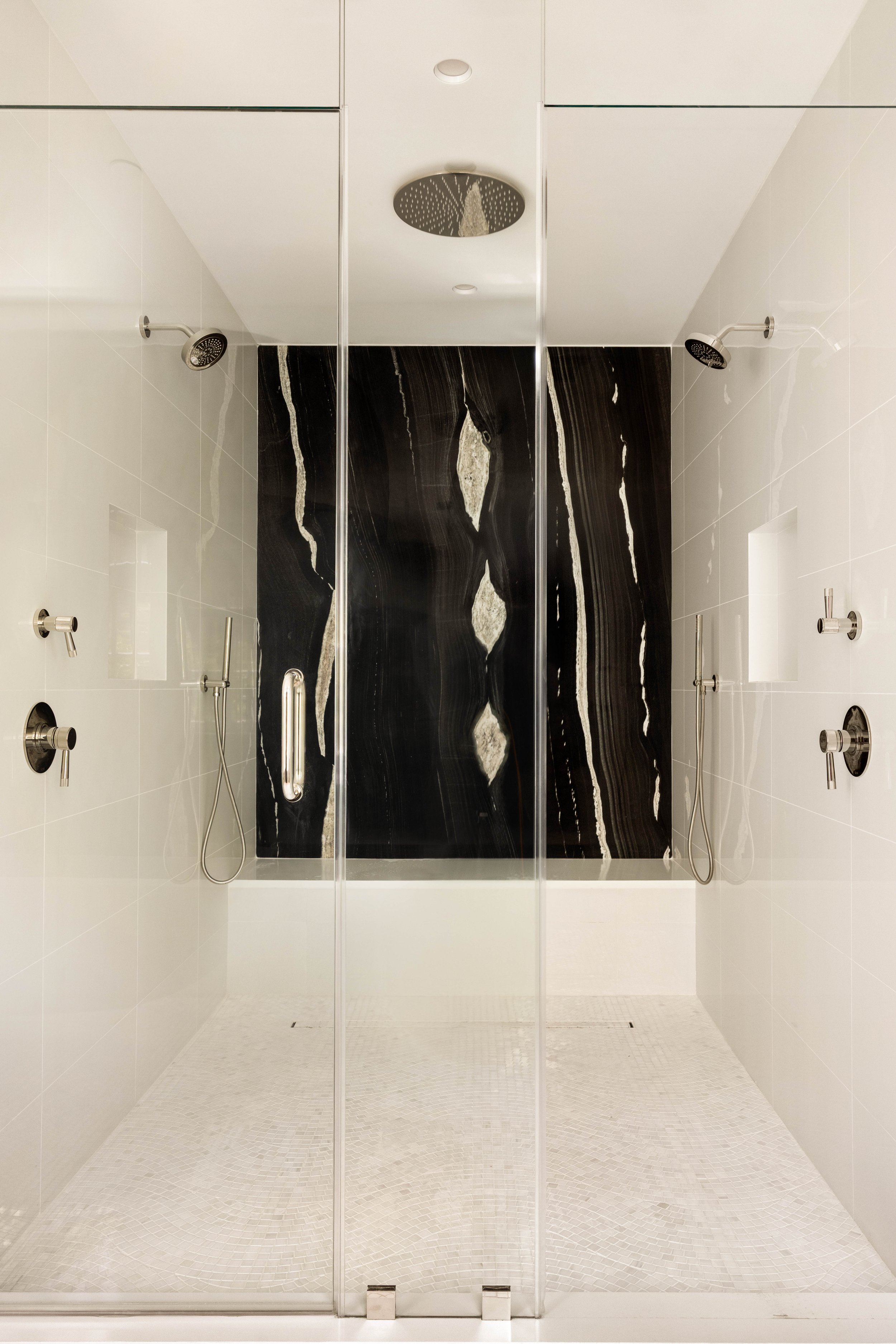 Modern shower with glass doors, black marble back wall, and six shower heads on white tiled walls.