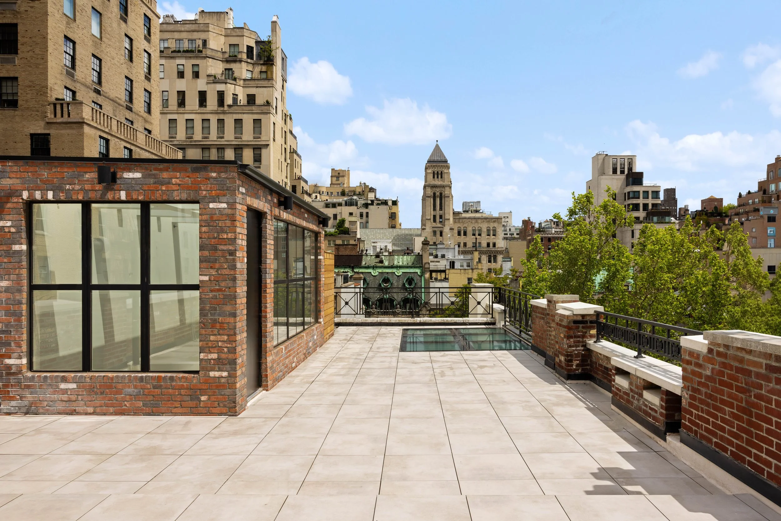 Rooftop terrace with beige tiled floor, brick walls, black metal railing, and cityscape view with historic buildings and blue sky with clouds.