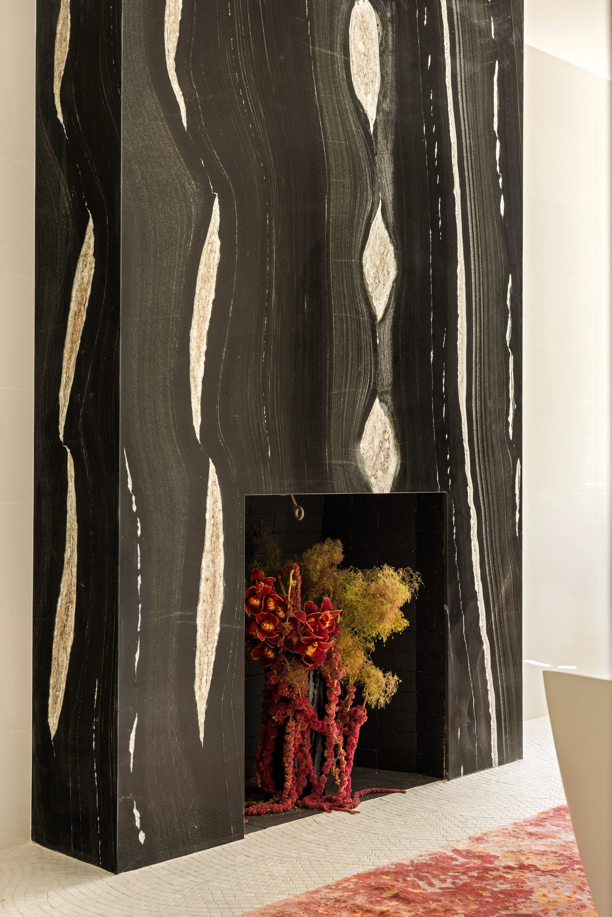 Interior space featuring a black marble wall with white and beige veining. There is a small built-in area with decorative red and green plants, partially visible at the bottom. The floor is tiled with small white tiles and a reddish patterned rug is 
