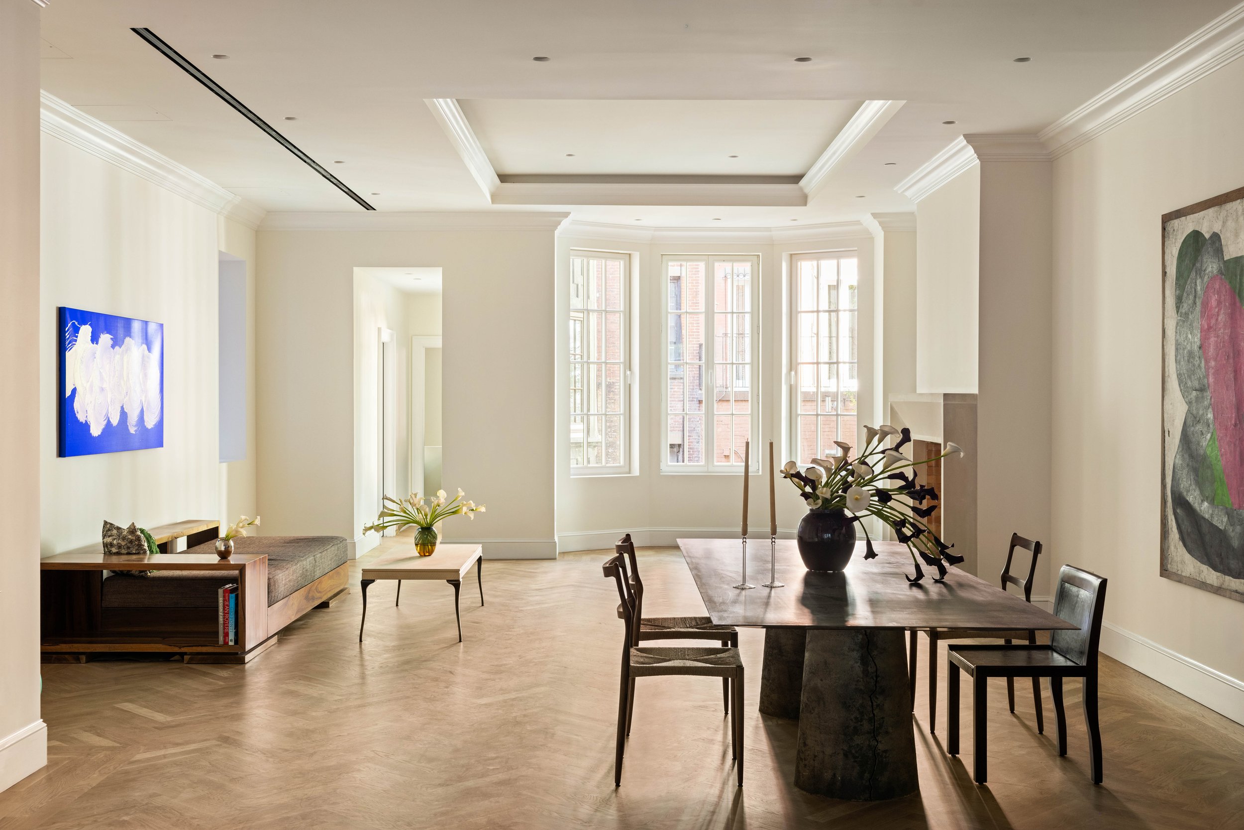 Living room with large bay windows, wooden dining table with chairs, a side table with books, and modern art on the walls.