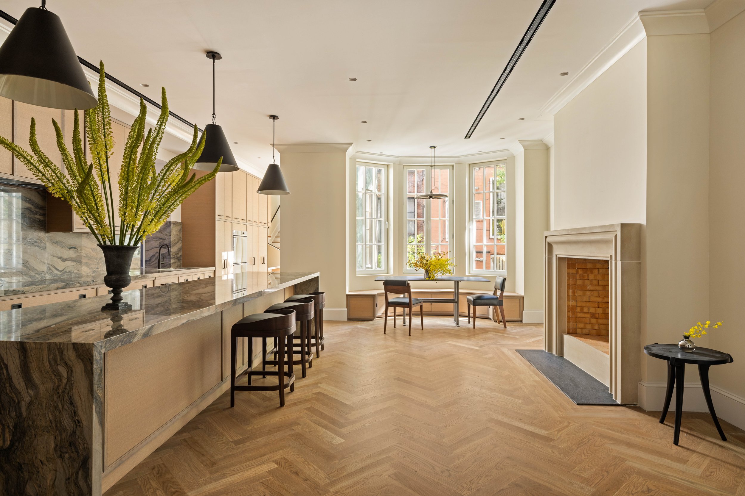 Modern kitchen and dining area with large bay windows, a marble kitchen island with bar stools, a fireplace, and a vase with tall green plants on the island.