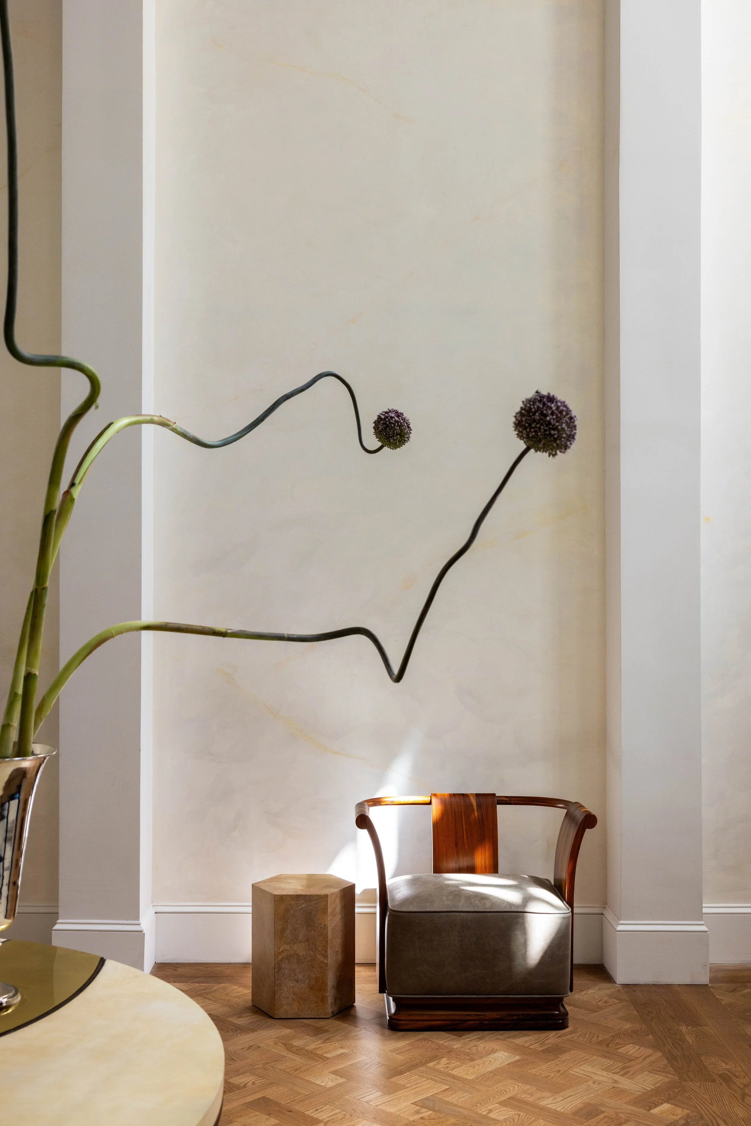 A modern, minimalist interior with a beige wall, a wooden chair with a light-colored cushion, a small wooden side table, a large tall vase with curvy dark branches and purple spherical flowers, and a hexagonal marble stool on a herringbone wood floor