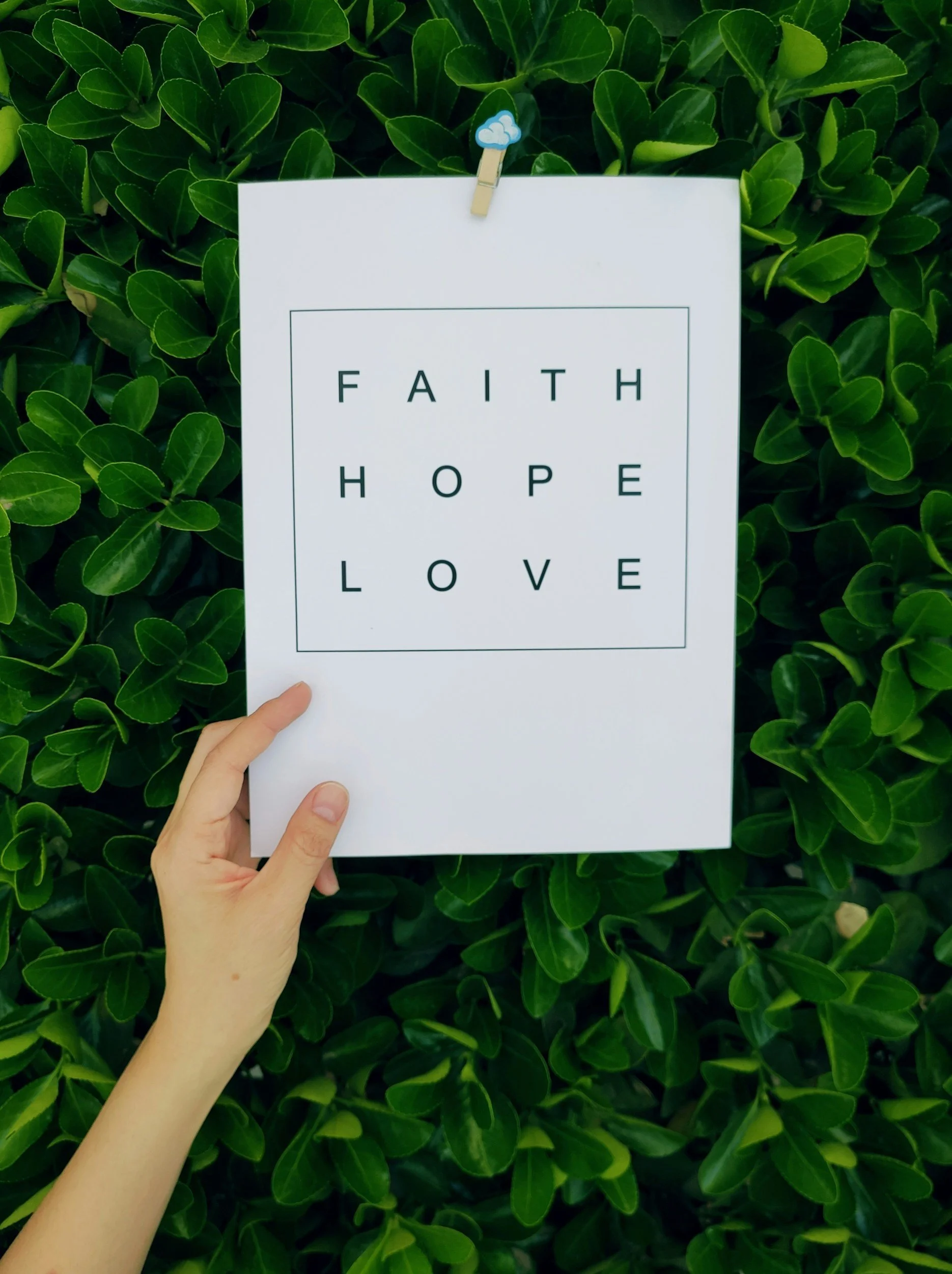 A person holding a white paper with the words 'FAITH HOPE LOVE' written on it against a background of green leaves.