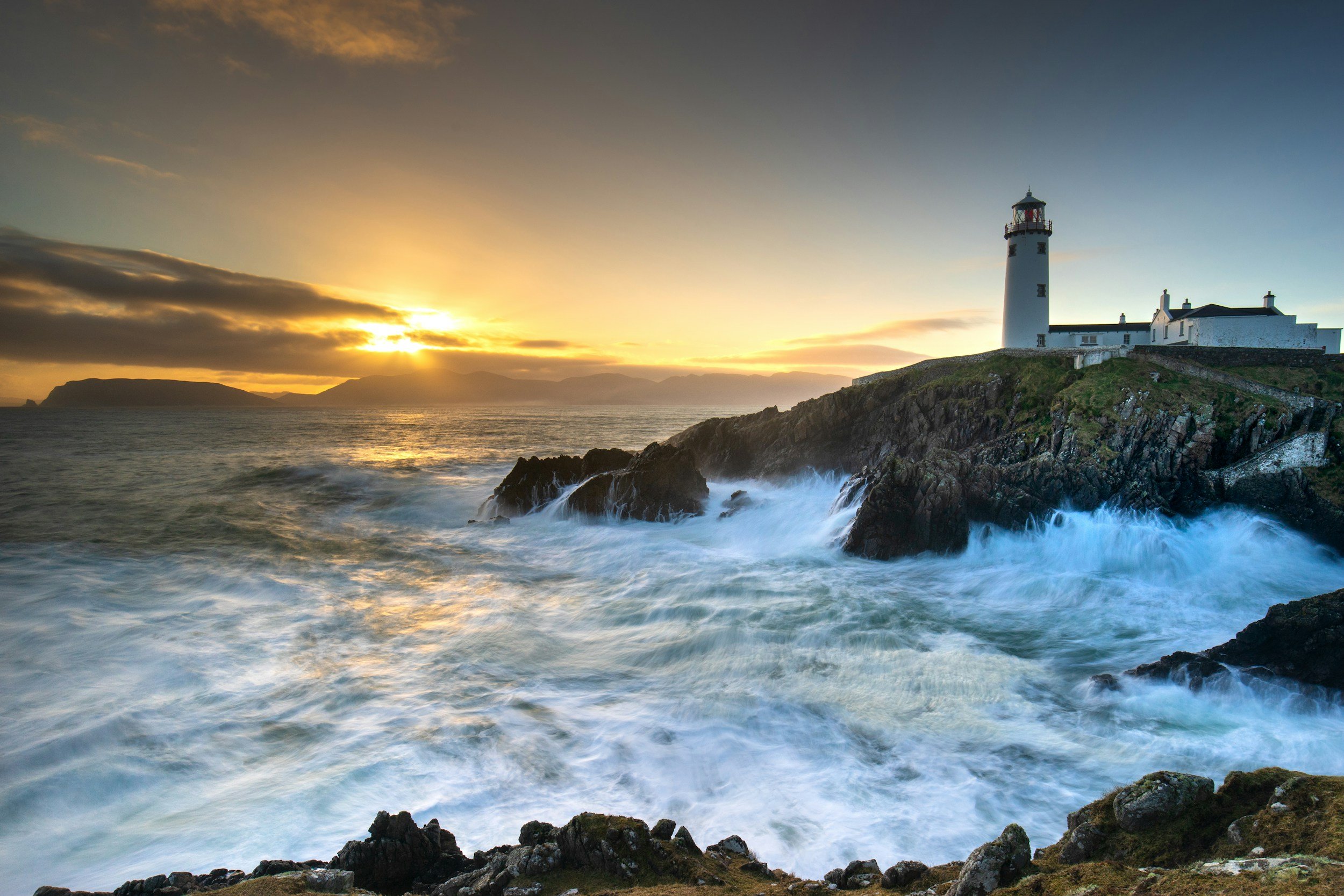 A lighthouse on a rocky coast with waves crashing, a sunset with clouds in the sky, and distant hills or islands.