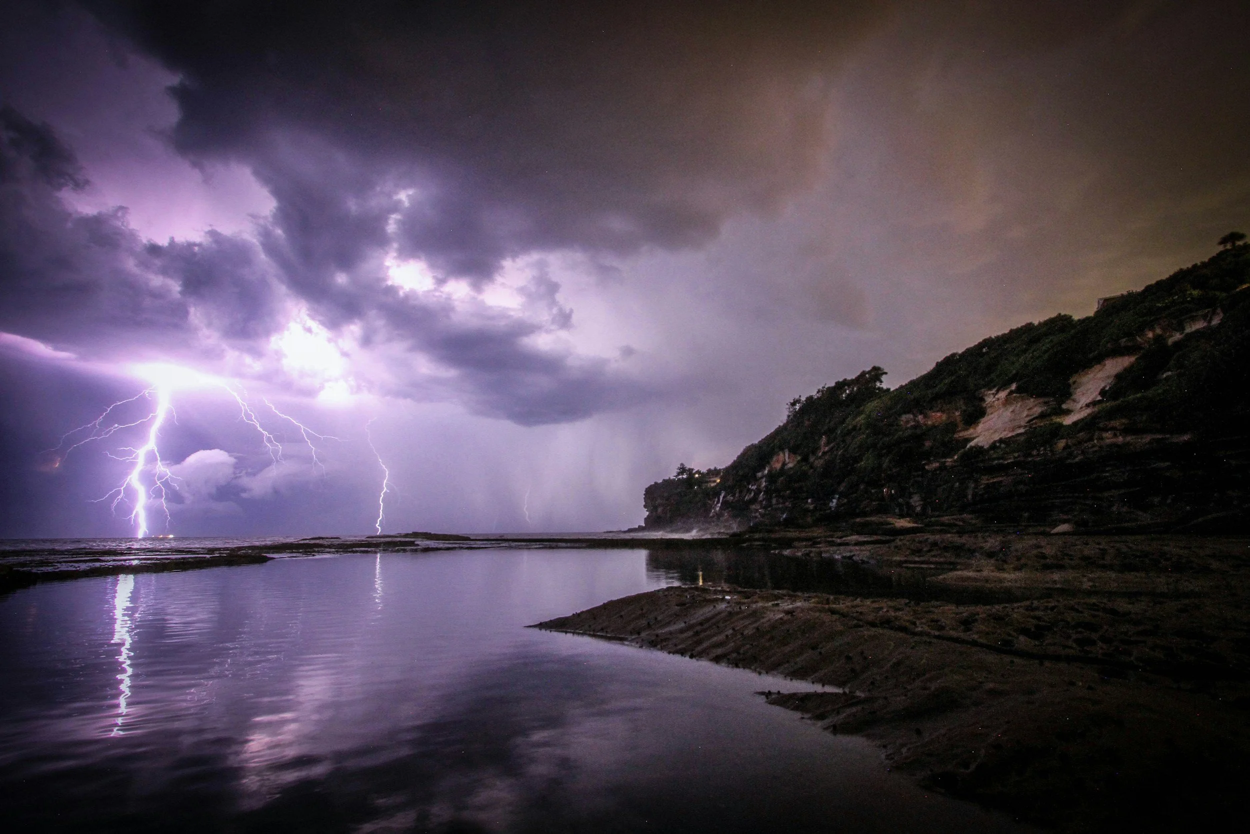 Lightning strikes over a dark, cloudy sky and reflects on a calm body of water near a rocky shoreline with trees on a hillside.