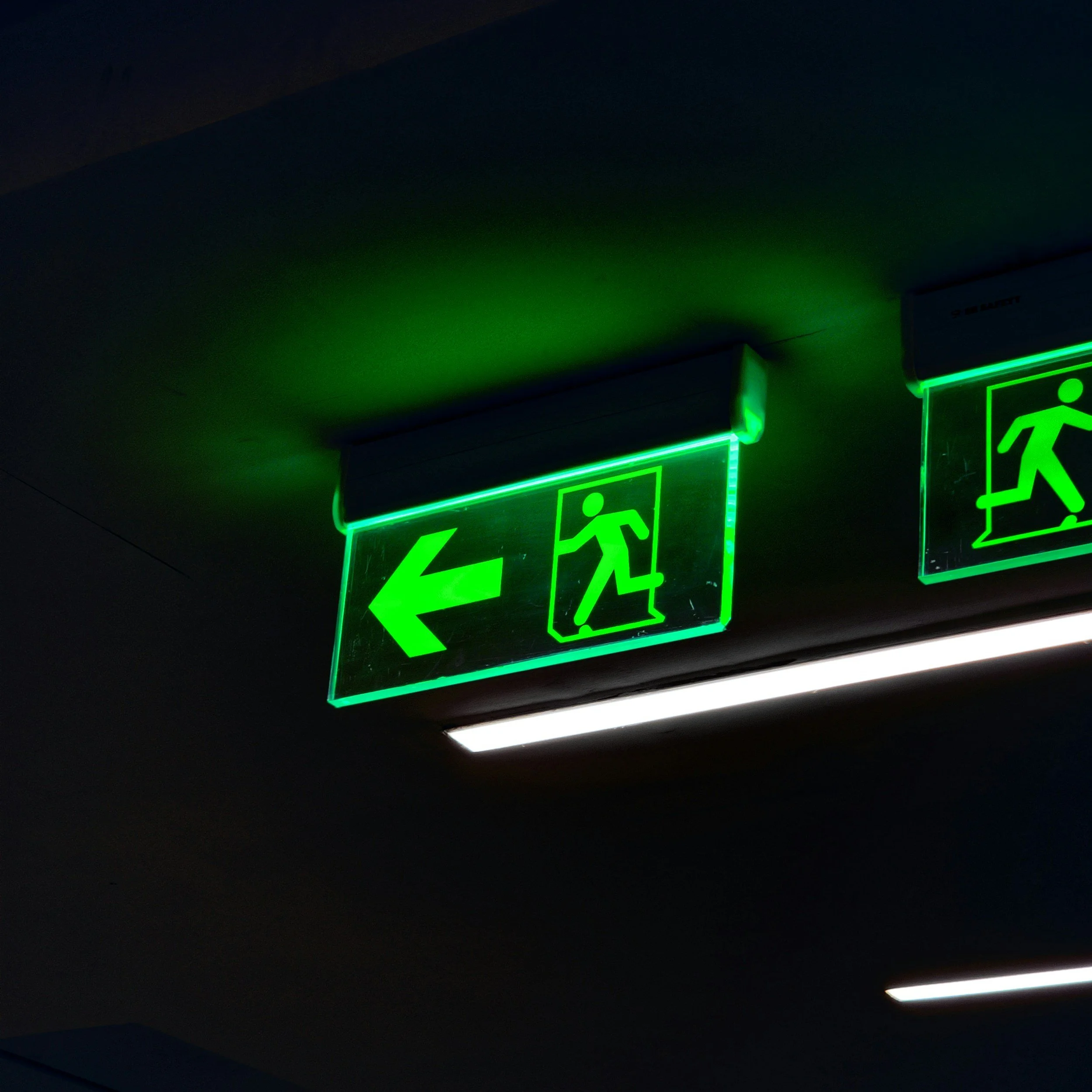 Green emergency exit sign with a running person symbol and an arrow pointing left, illuminated with neon lighting.
