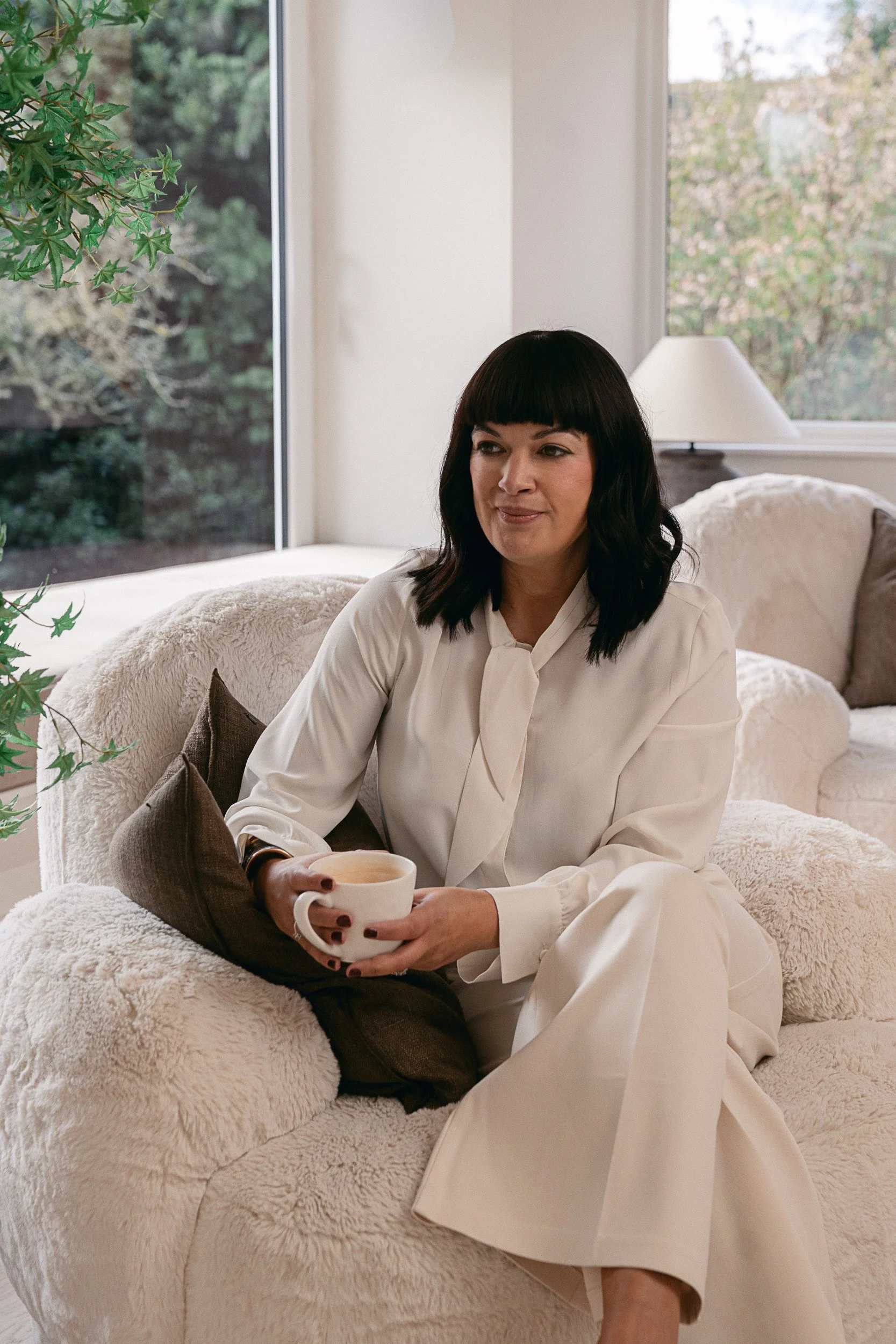 A woman with dark hair and bangs sitting on a plush cream-colored sofa, holding a white mug, in a room with large windows showing trees outside, and a white table lamp in the background.