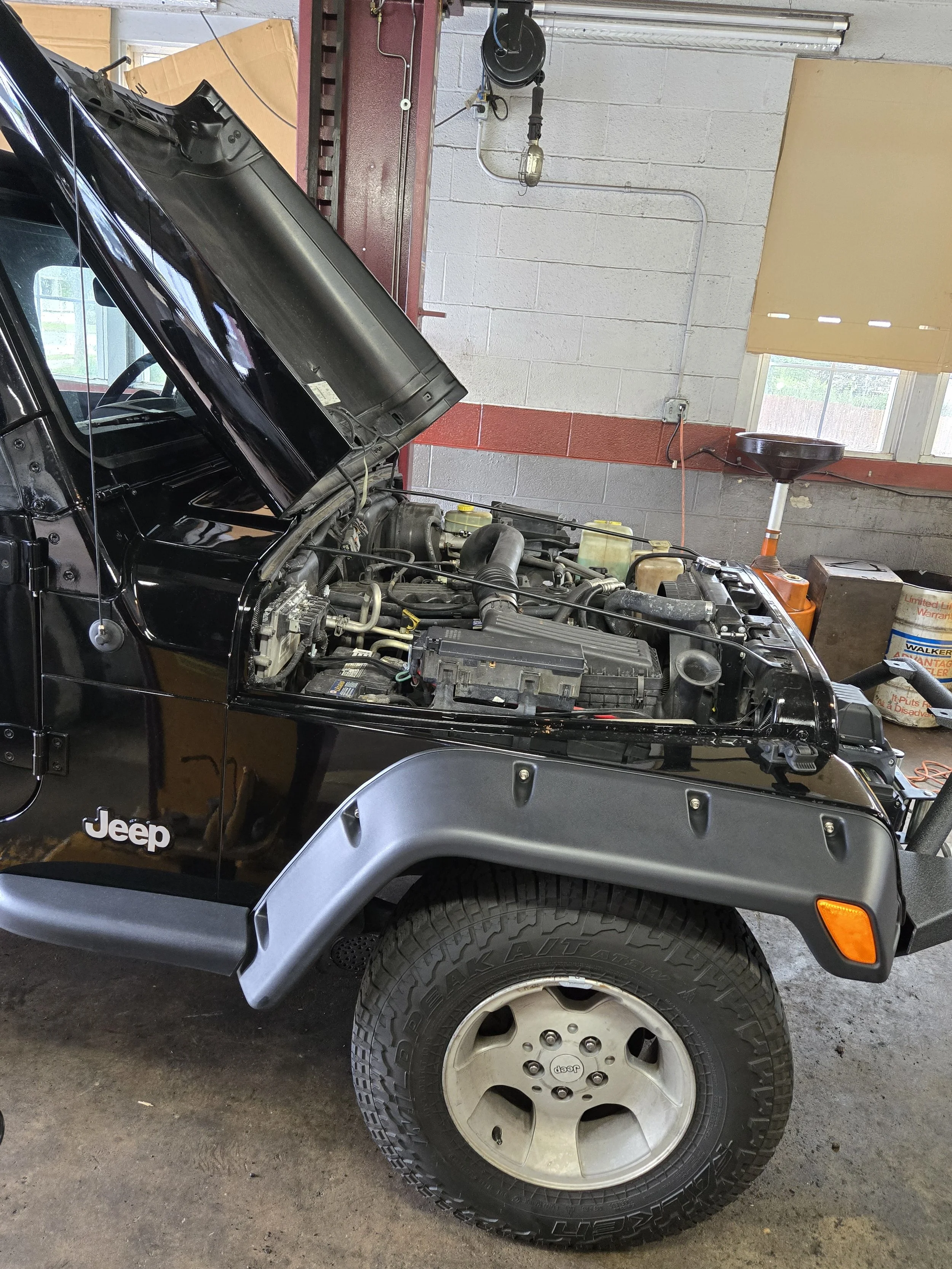 A black Jeep with its hood open inside a garage, showing the engine. The front right wheel of the Jeep is visible, and the background includes a window, a stool, and various tools and equipment.