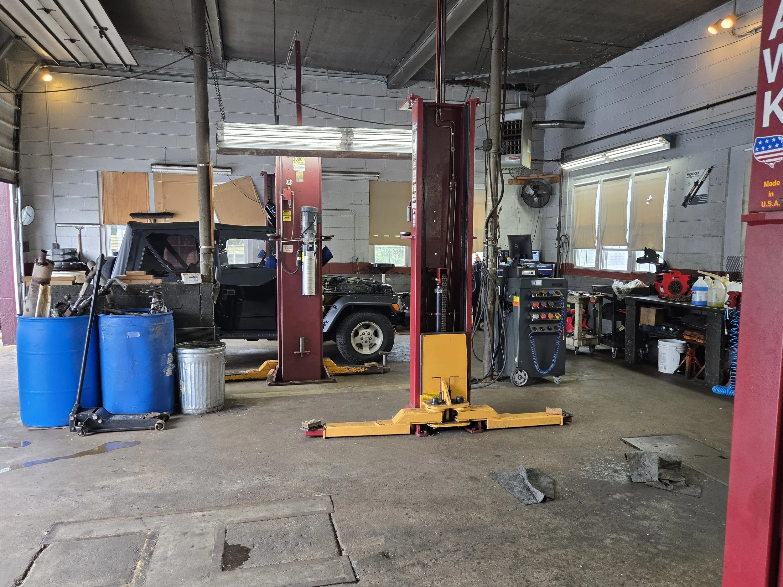 Inside an auto repair shop with a car on a hydraulic lift, blue barrels, and various tools and equipment scattered around.