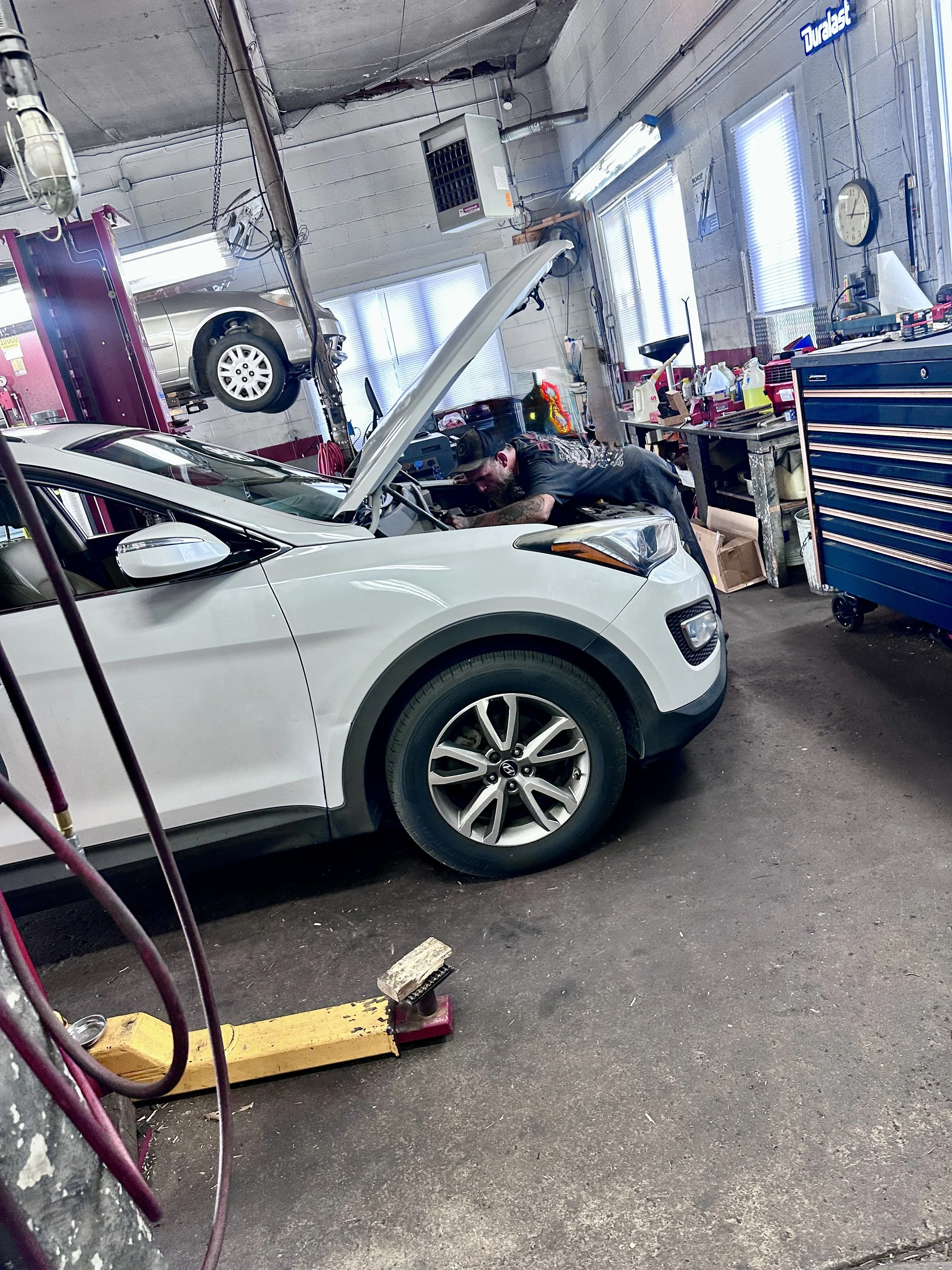 An auto repair shop with a white SUV on the garage floor, a mechanic working under the open hood, tools and supplies on workbenches, a car lift with another vehicle elevated, and various equipment and equipment in the background.