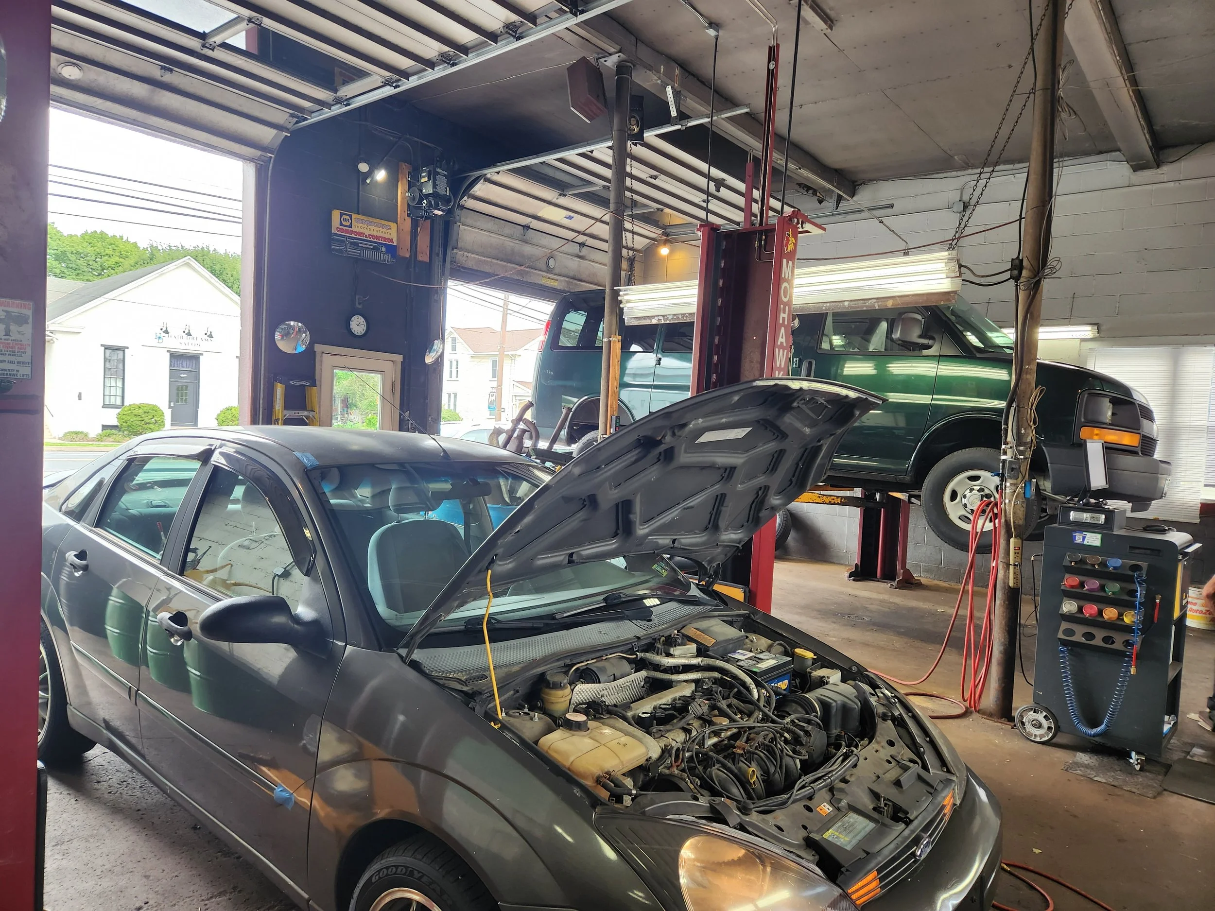 An automotive repair shop with a silver car in the foreground having its hood open, and a green truck on a lift in the background. The shop has tools, equipment, and fluorescent lighting.