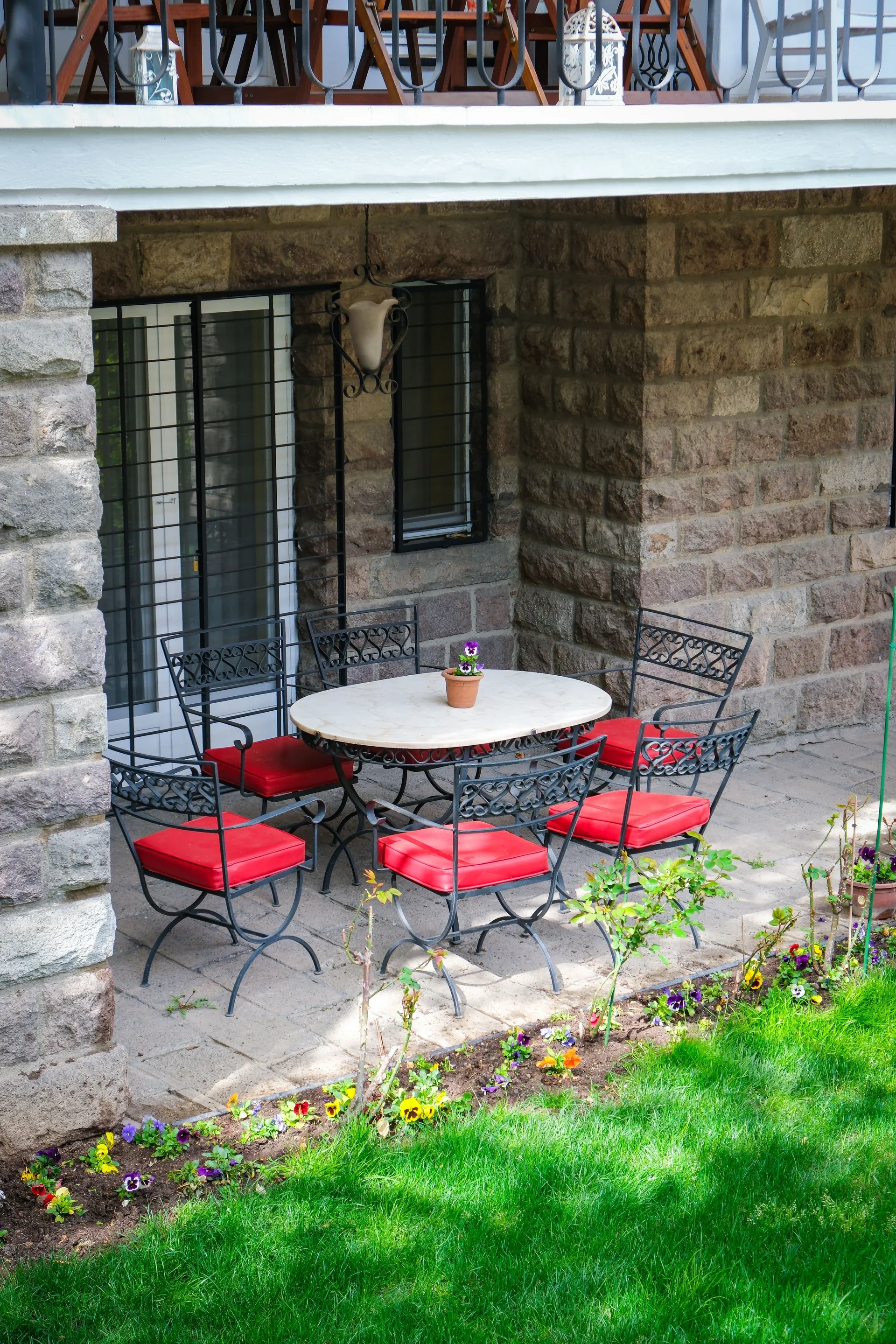 A cozy outdoor patio area with a white marble table, surrounded by black wrought iron chairs with red cushions. A small potted purple flower is on the table. There is a stone wall with a window and window bars behind the table, and a decorative wall lantern hanging above. The patio floor is made of stone tiles, and there are colorful flowers and green grass in the foreground.