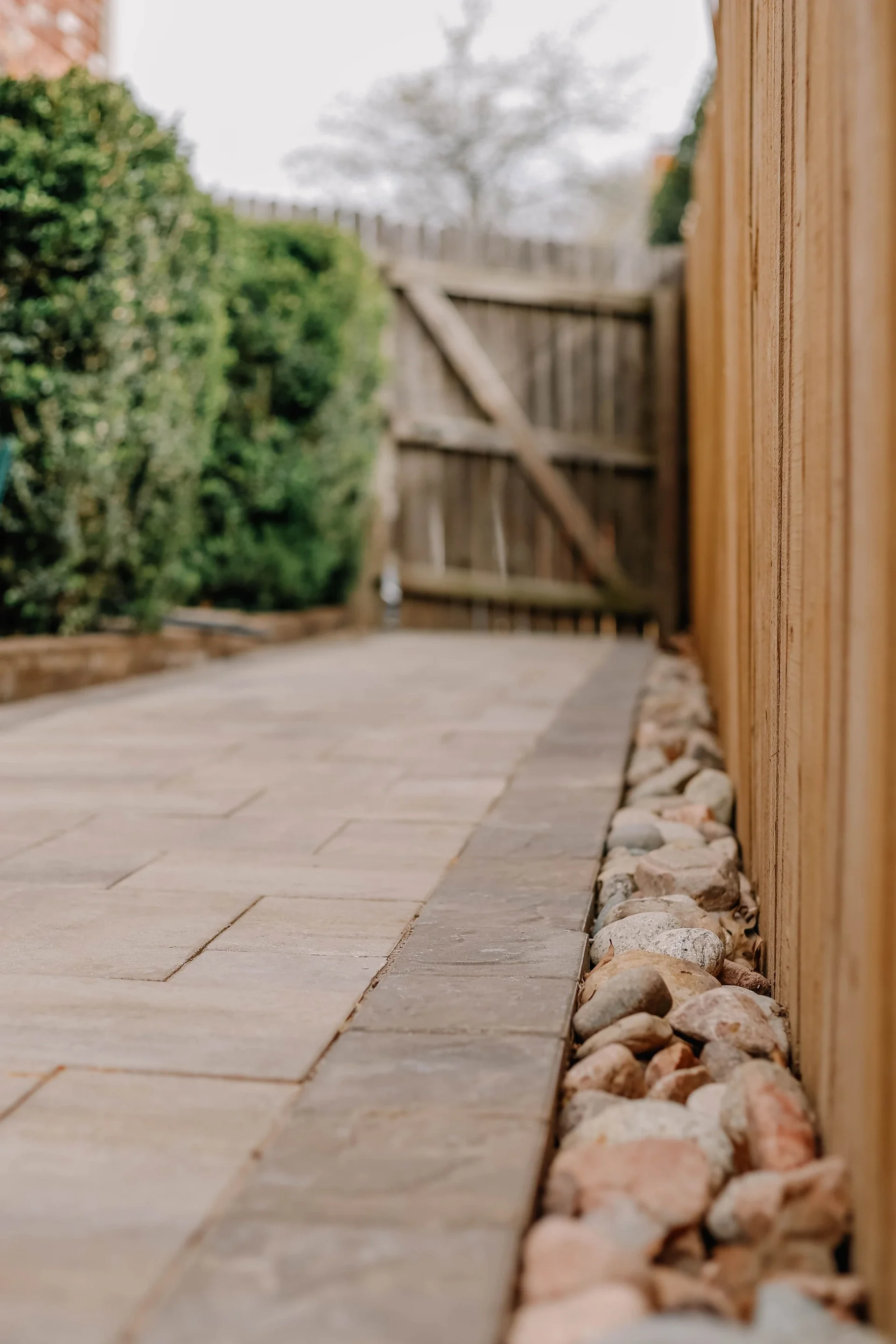 A close-up view of a side yard with a wooden fence on the right, a paved walkway, a row of small rocks lining the fence, and greenery including a hedge and a tree in the background.