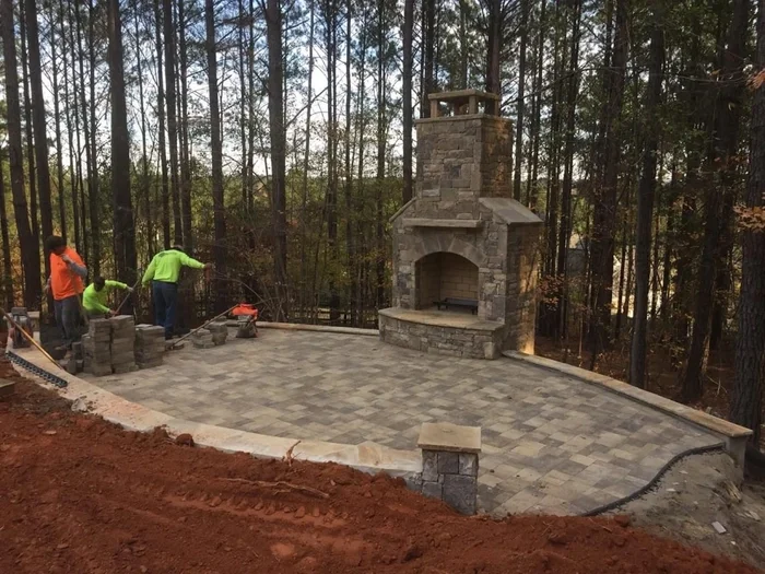 Workers are building a paved outdoor patio with a stone fireplace in a wooded area.