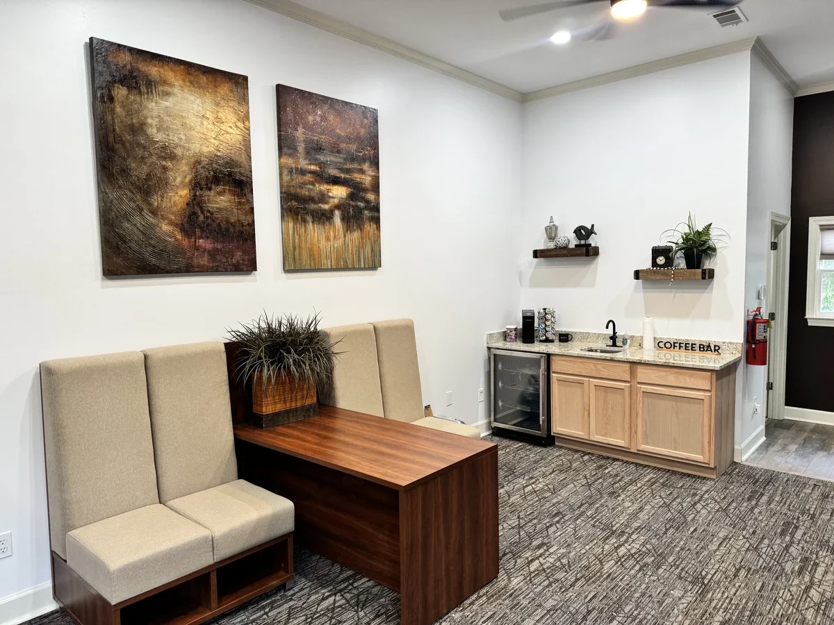 Interior of a small hospitality or lounge area with a seating booth, a table with a plant, wall art, a mini fridge, a coffee maker, and shelves with decorative items.