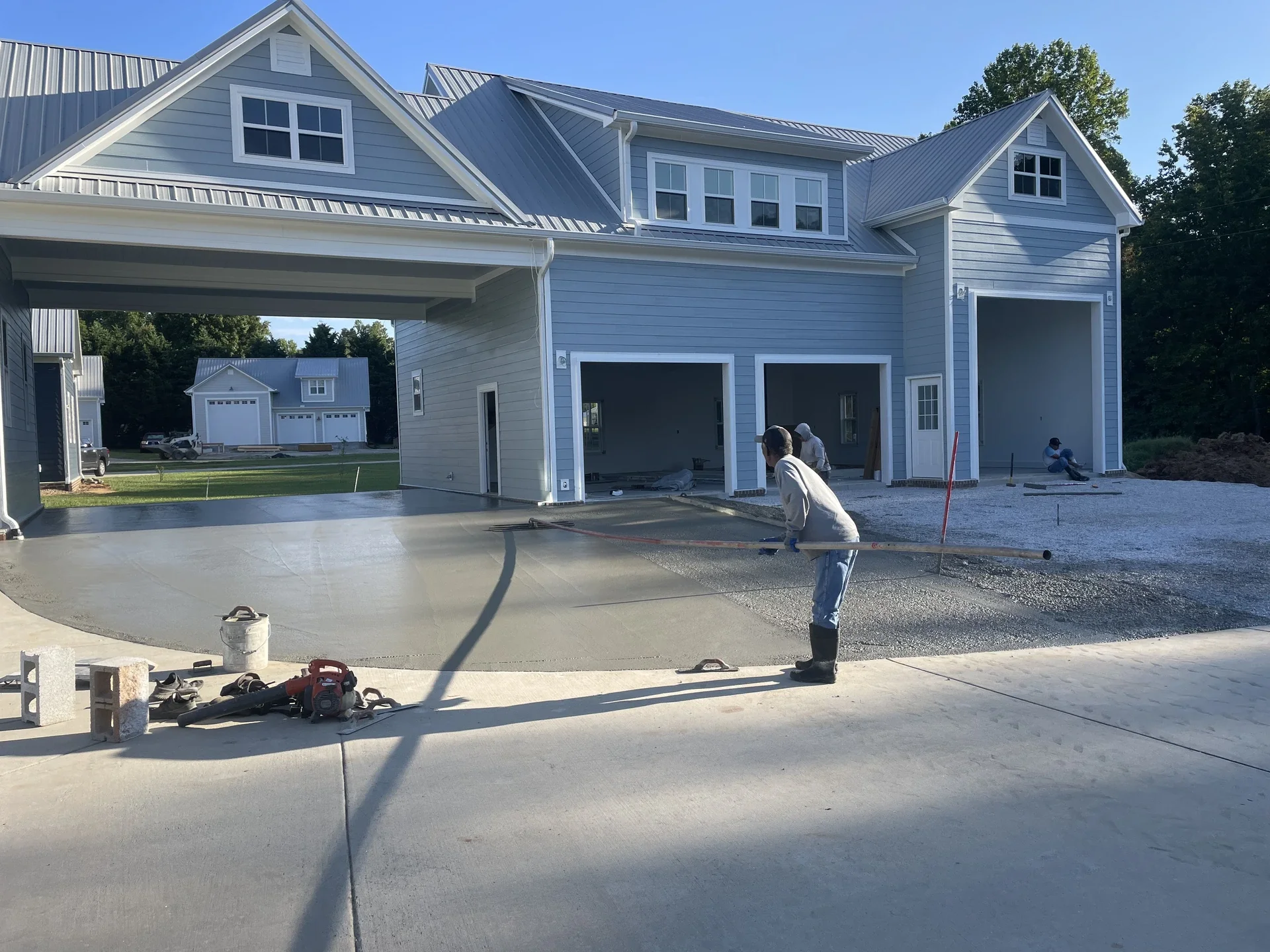 Construction workers pouring and spreading concrete for a driveway or patio in front of a light blue house with white trim. Several construction tools and materials are in the foreground.