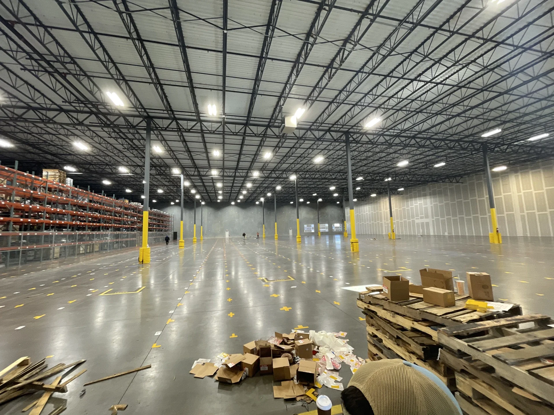 Empty warehouse with high ceiling, metal beams, bright overhead lights, and scattered cardboard boxes and pallets on the floor.