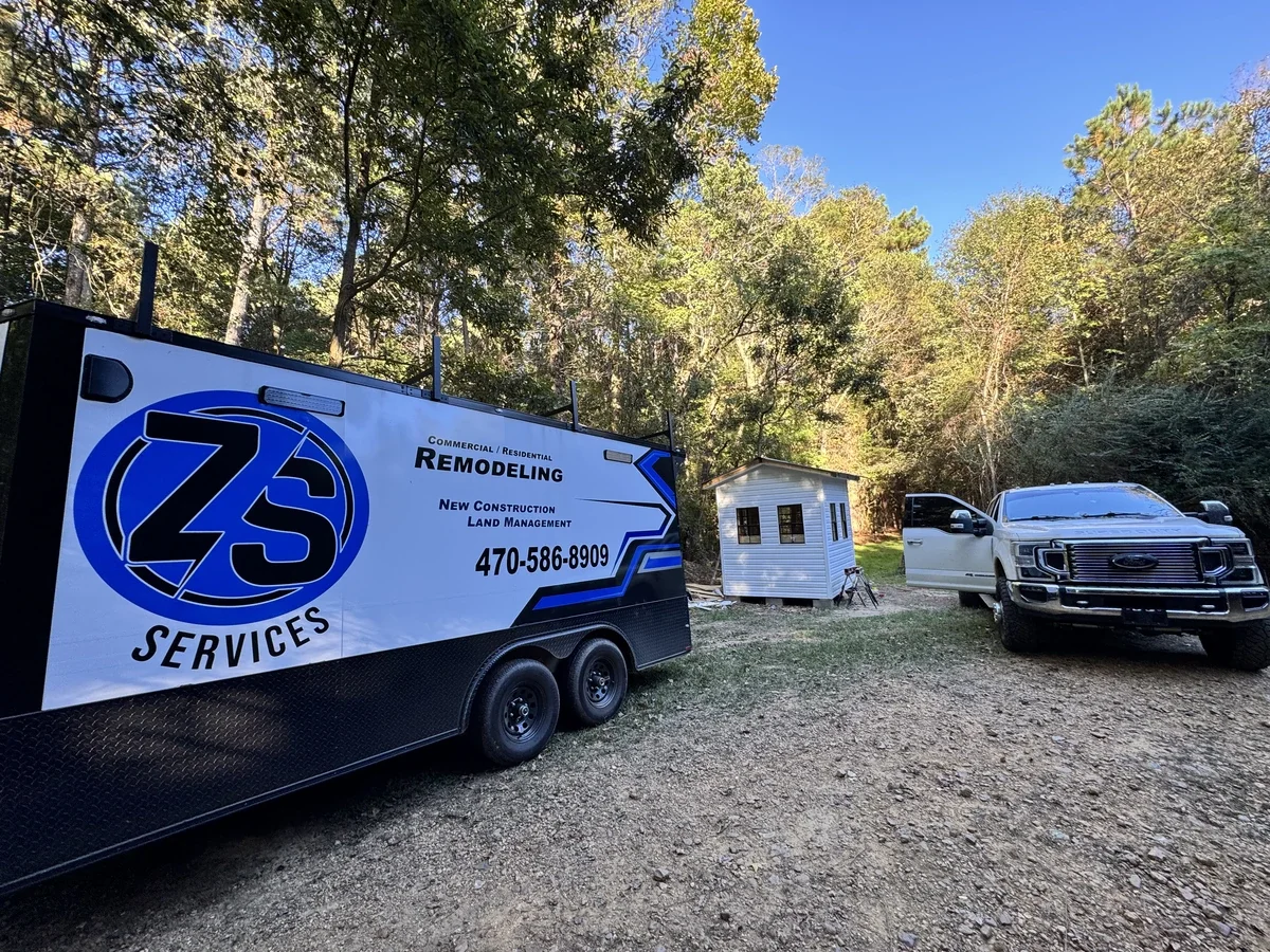 ZS Services trailer, a white truck with an open door, and a small white building in a wooded area.