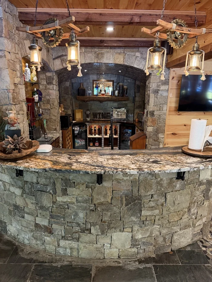 A rustic home bar with a stone curved counter, wooden accents, hanging lantern-style lights, and a stone arched opening leading to a storage area with bottles of alcohol.