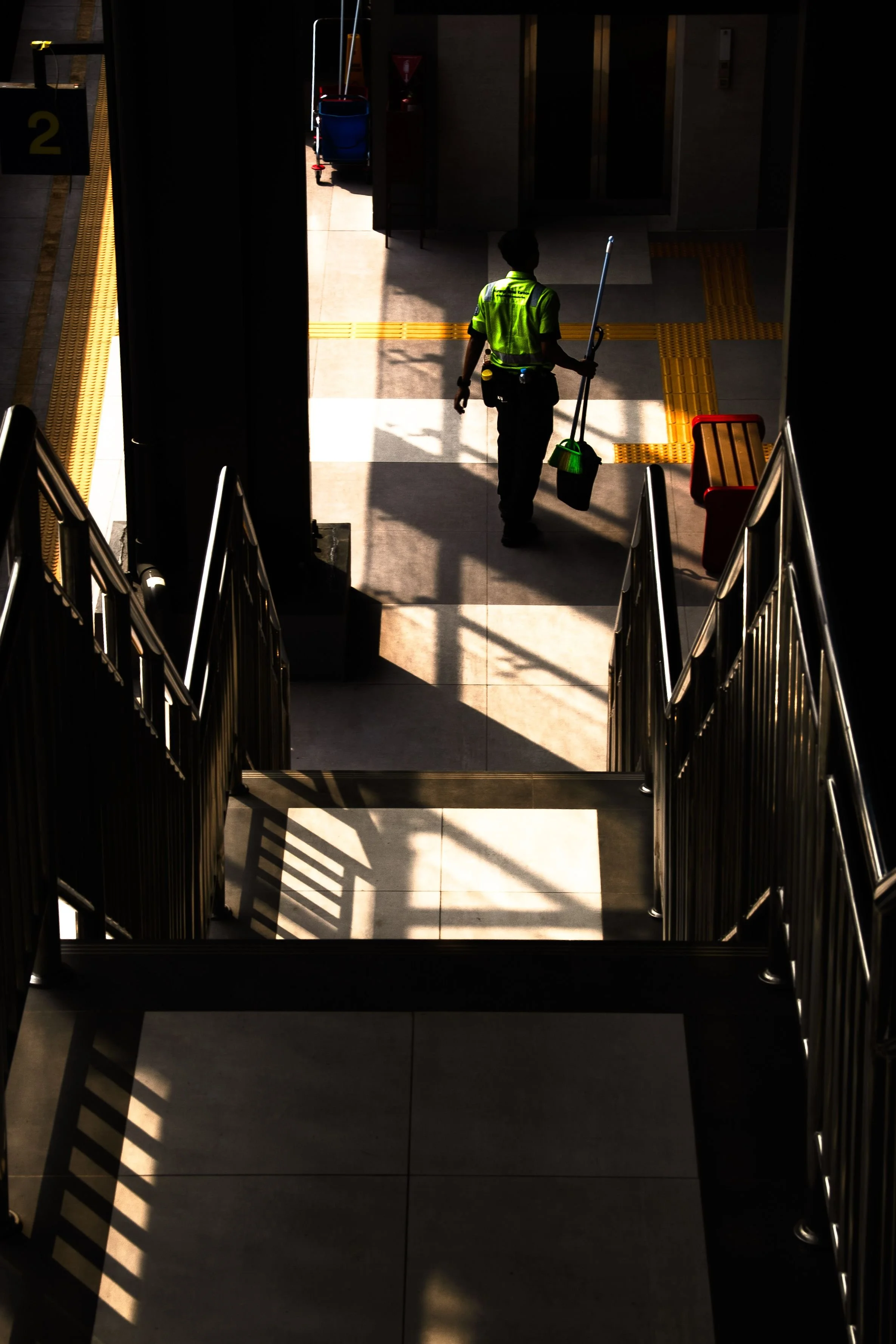 A person in a yellow safety vest walking down a sunlit corridor with railings on the sides, carrying a mop and bucket, in what appears to be a commercial or public building.