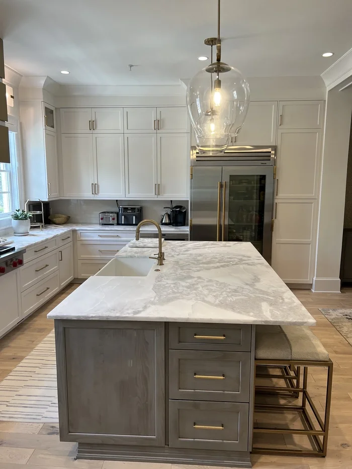 Modern kitchen with white cabinets, a marble island with an under-mount sink, and a stainless steel refrigerator. A pendant light hangs above the island.