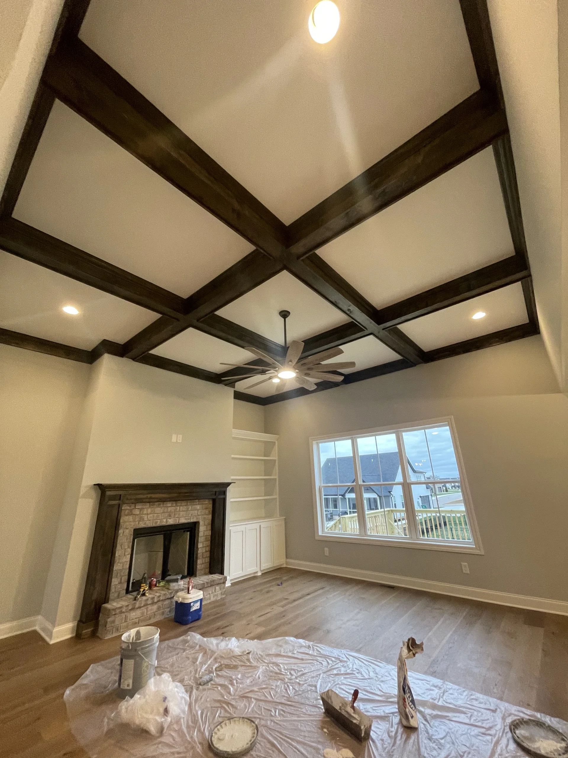 Living room with wood beam ceiling, window, fireplace, and ceiling fan, under renovation with painter's tarp and painting supplies on the floor.