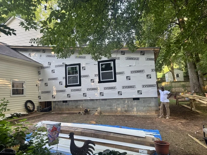 Back of a house under construction, with New vinyl siding and three windows installed on the upper part, surrounded by trees and construction materials on the ground.