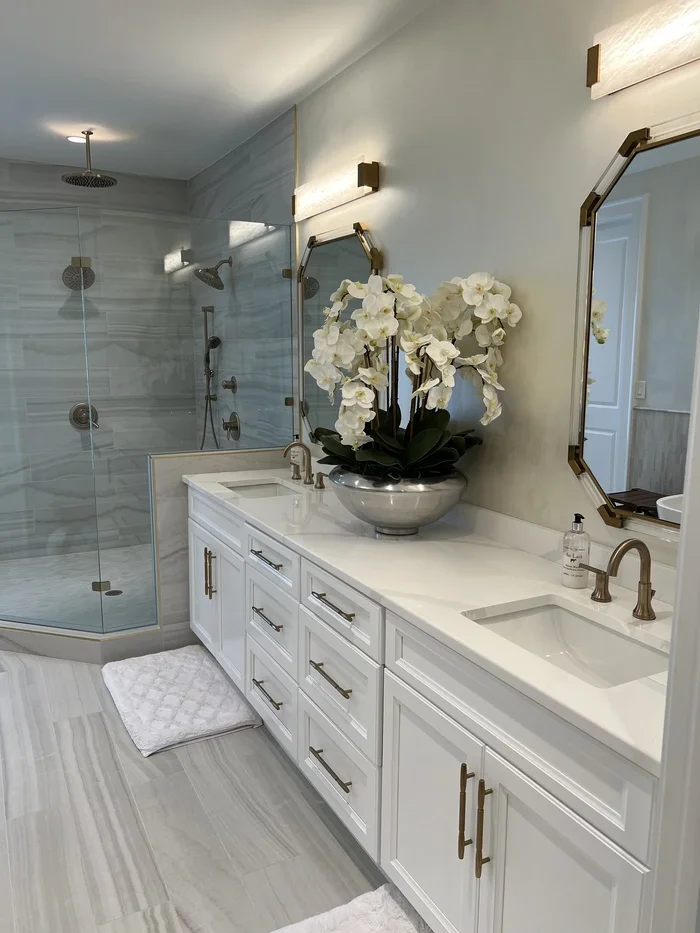 Modern bathroom with double vanity, white cabinets, gold handles, large mirror, white orchid plant, glass shower with multiple showerheads, and light gray tiled floor.