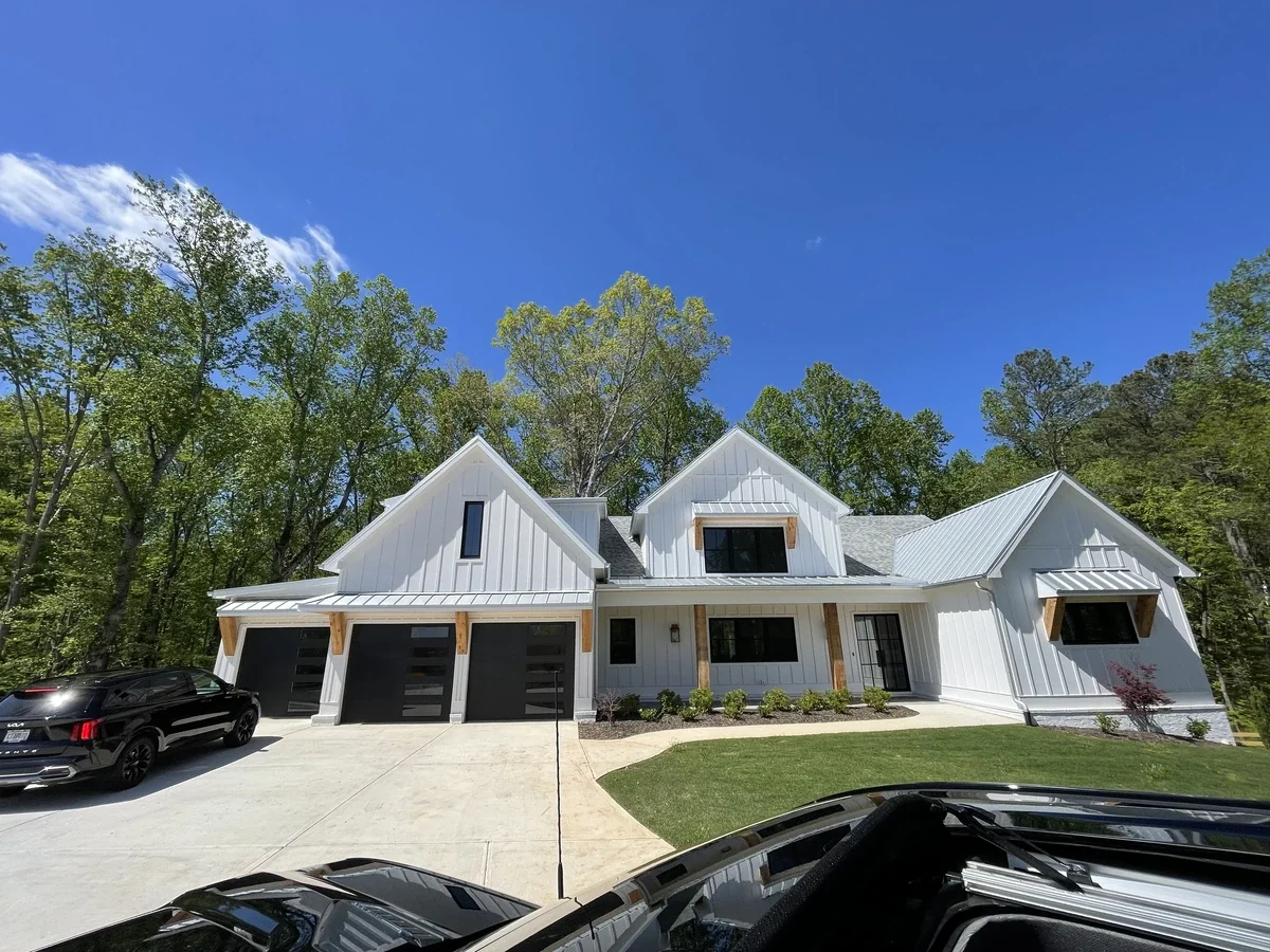 Modern white house with black garage doors and a driveway, surrounded by green trees under a blue sky.