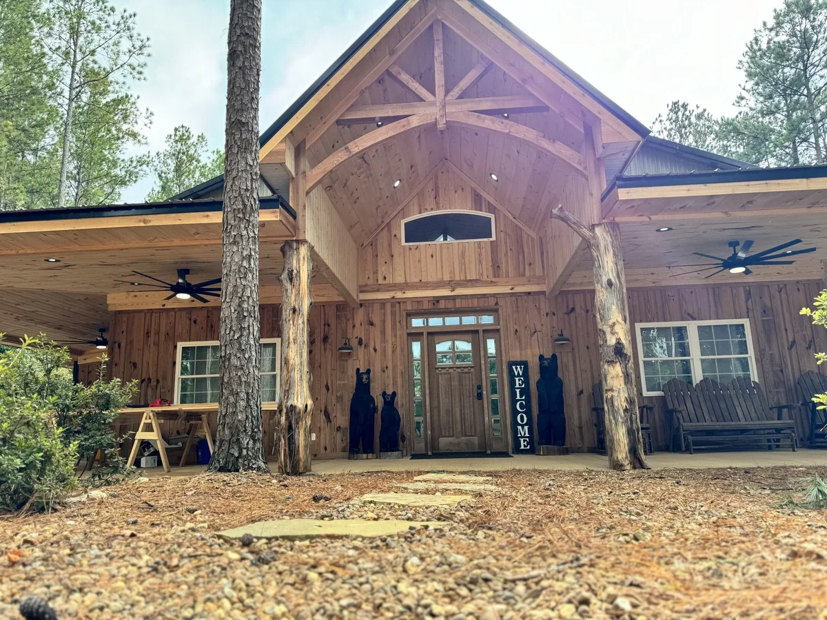 The front entrance of a rustic wooden house with a high peaked roof, surrounded by trees. There are two black bear sculptures and a welcome sign near the doorway, with an outdoor seating area and ceiling fans visible under the porch.