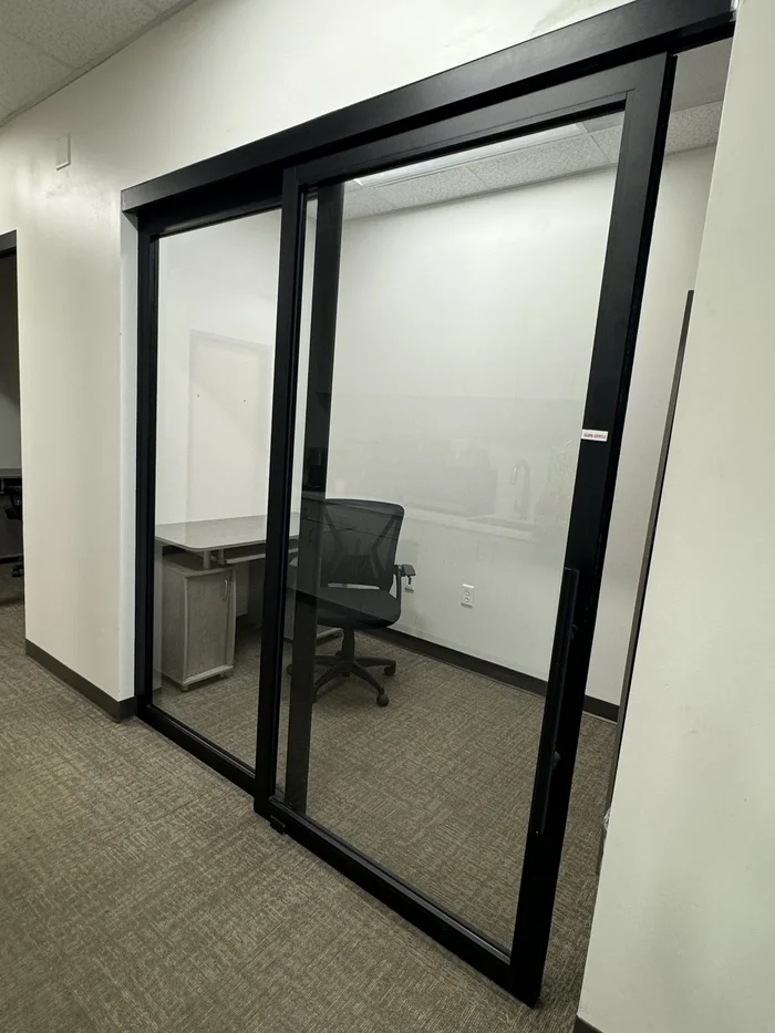 An office with a glass sliding door, a desk with a monitor, and an office chair inside the enclosed space.