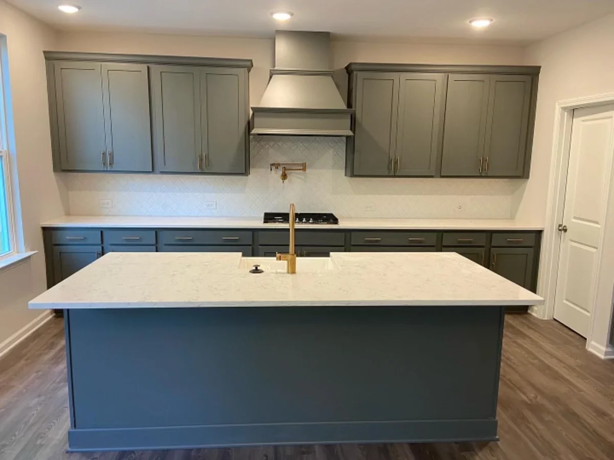 A modern kitchen with gray cabinets, a white countertop island, and a gold faucet. There are upper and lower cabinets with a range hood and stove in the middle. Recessed lighting and a window are on the left side, with a door on the right and wood flooring.