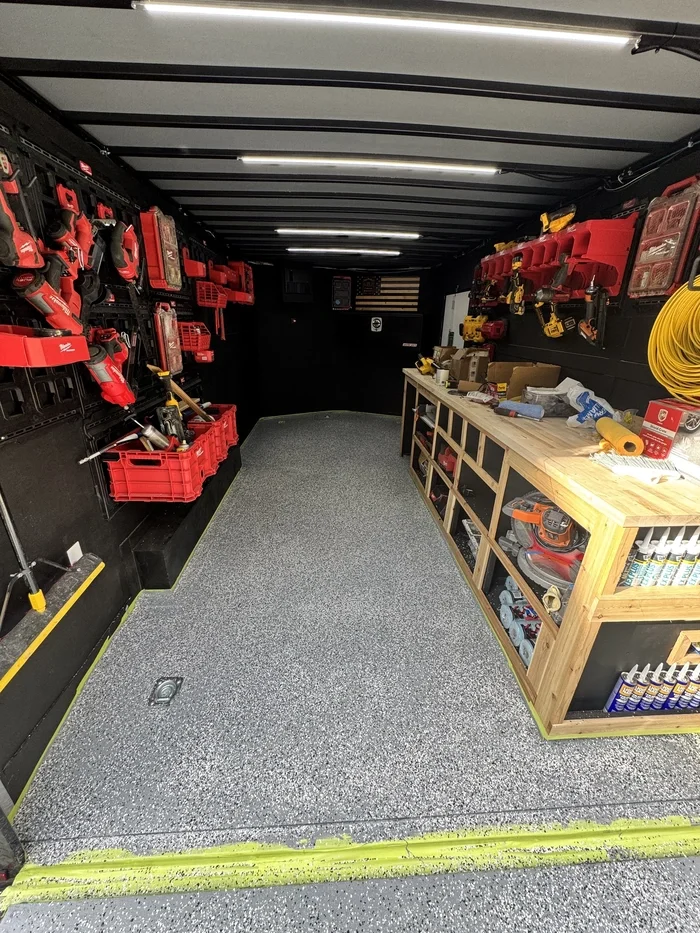 Tool storage area in a van, with red and yellow power tools on black shelves and a wooden workbench with various supplies.