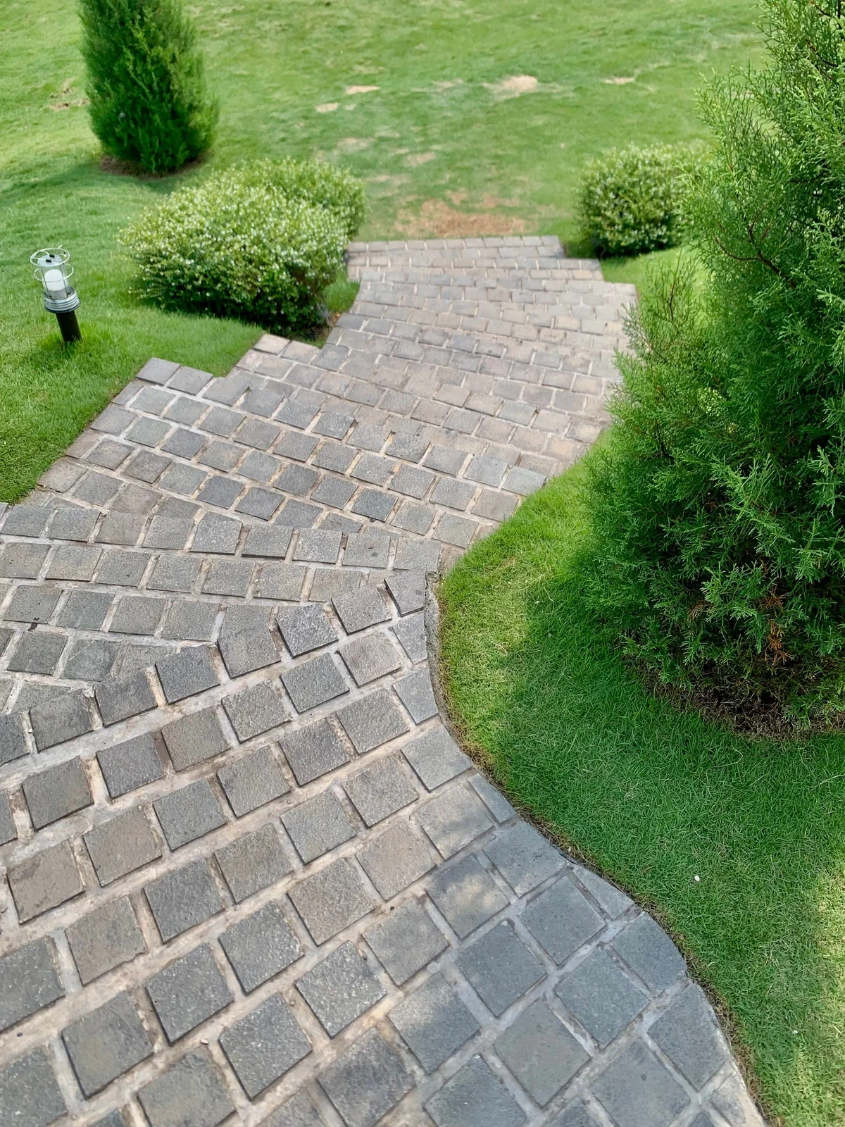 A paved walkway made of gray bricks curves through a well-manicured green lawn with bushes and small trees, and a small lantern-style garden light on a black post on the left side.