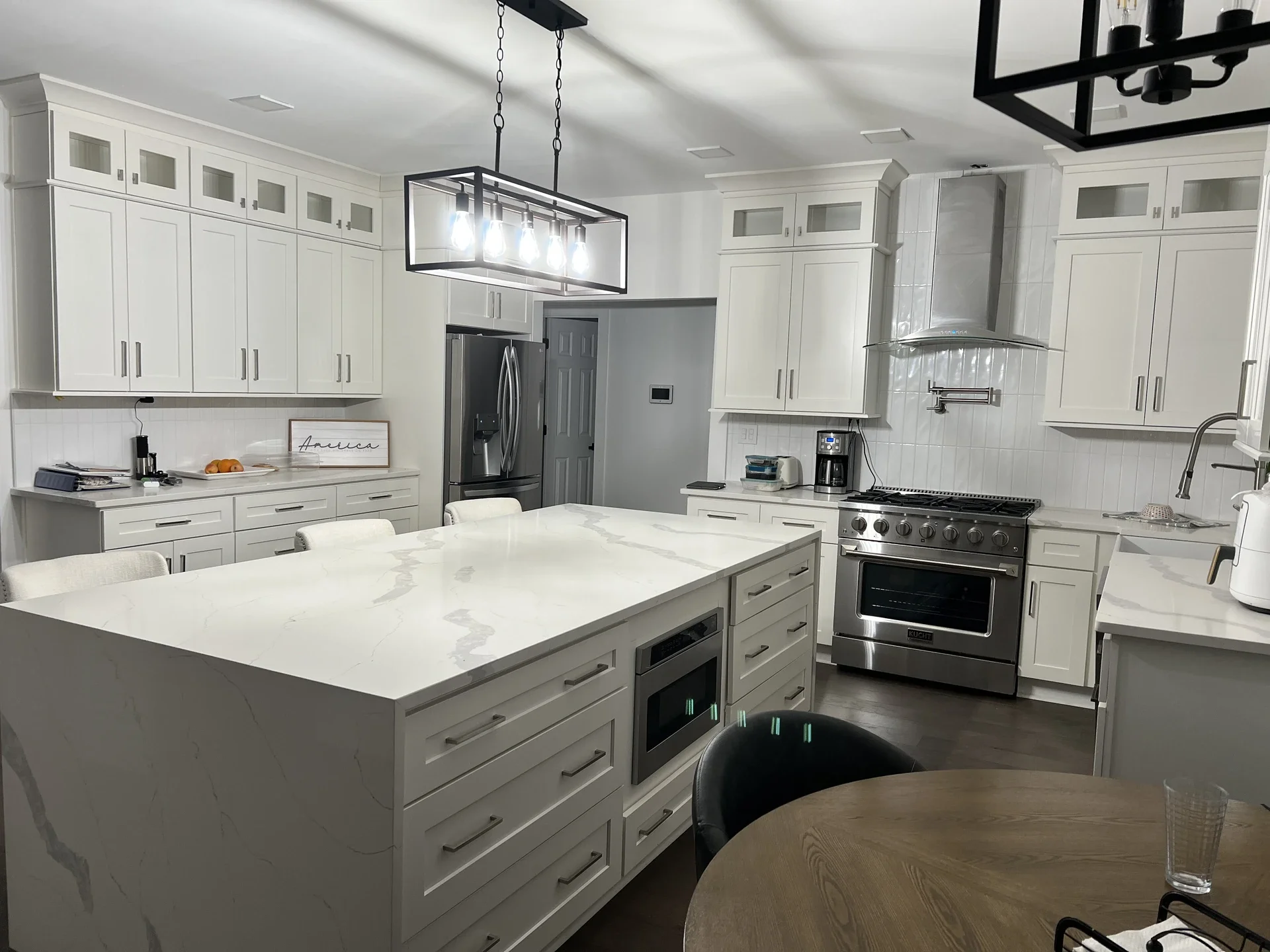 Modern kitchen with white cabinets, a large marble island, stainless steel appliances, a black dining chair, and a wooden dining table with a glass of water.
