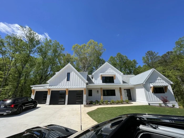 Modern white house with multiple gabled roofs, adjacent to a driveway with two parked cars, surrounded by green trees and a clear blue sky.