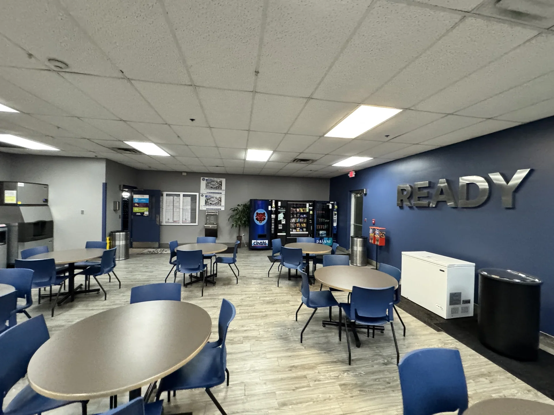 A break room or lounge with round tables and blue chairs, vending machines, a bulletin board, and a wall with the word "READY" in large metallic letters.