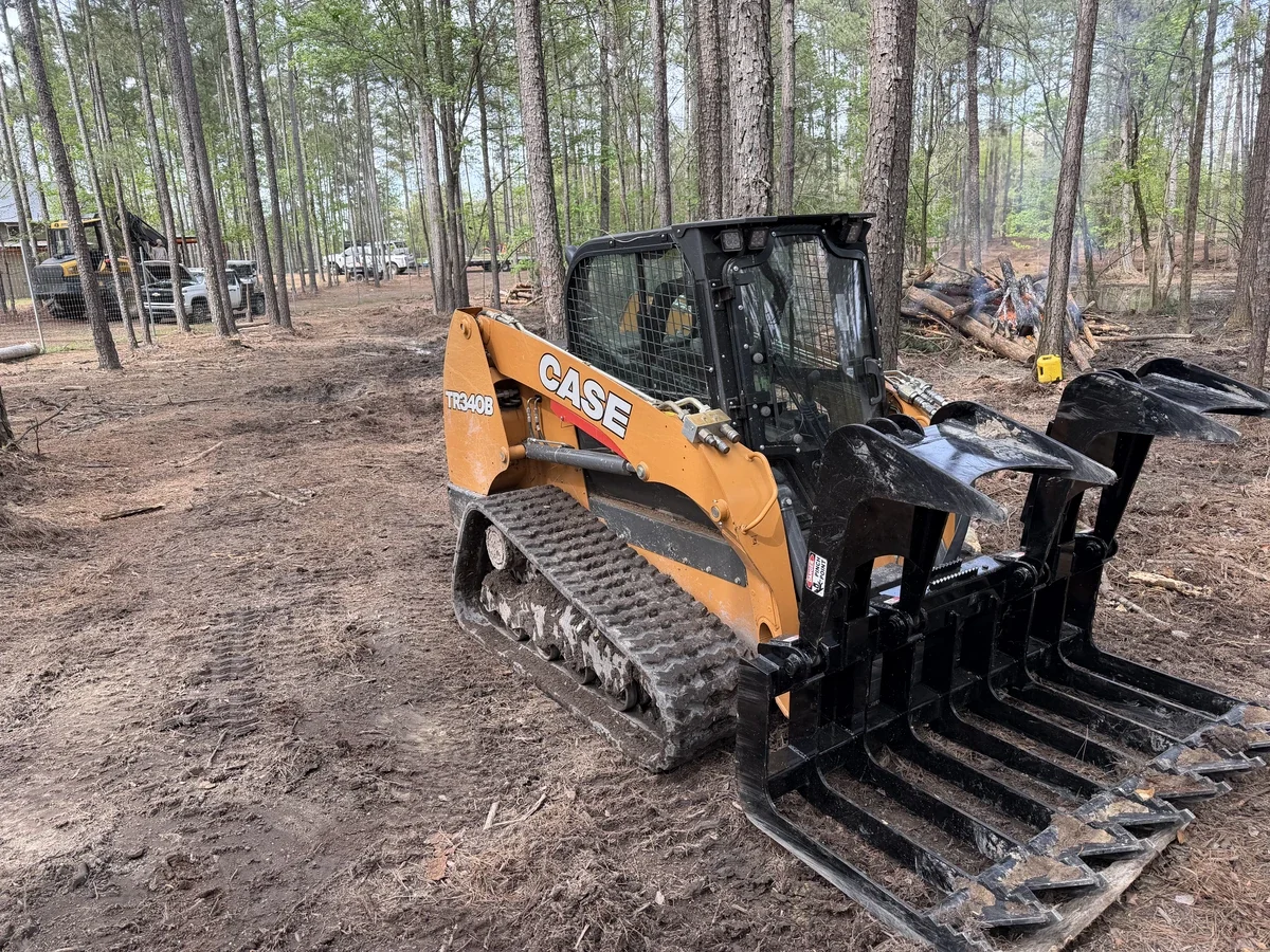 A CASE skid steer loader with a grapple attachment parked on a dirt clearing in a forested area, with trees and some vehicles in the background, and a small fire with logs burning nearby.