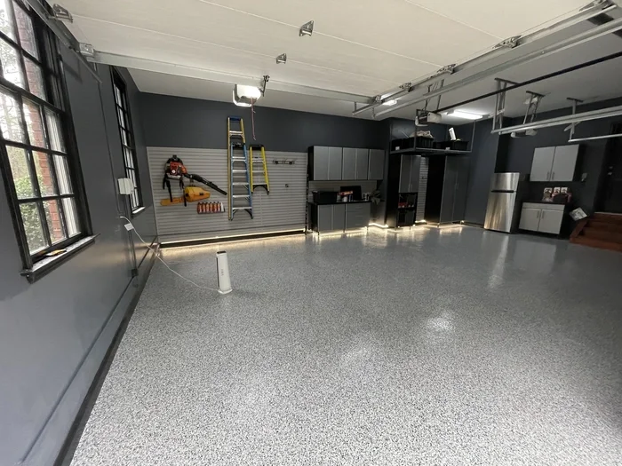 Empty garage with gray walls, open windows, grey cabinets, a small kitchenette, a toolbox, a ladder, and a stainless steel refrigerator.